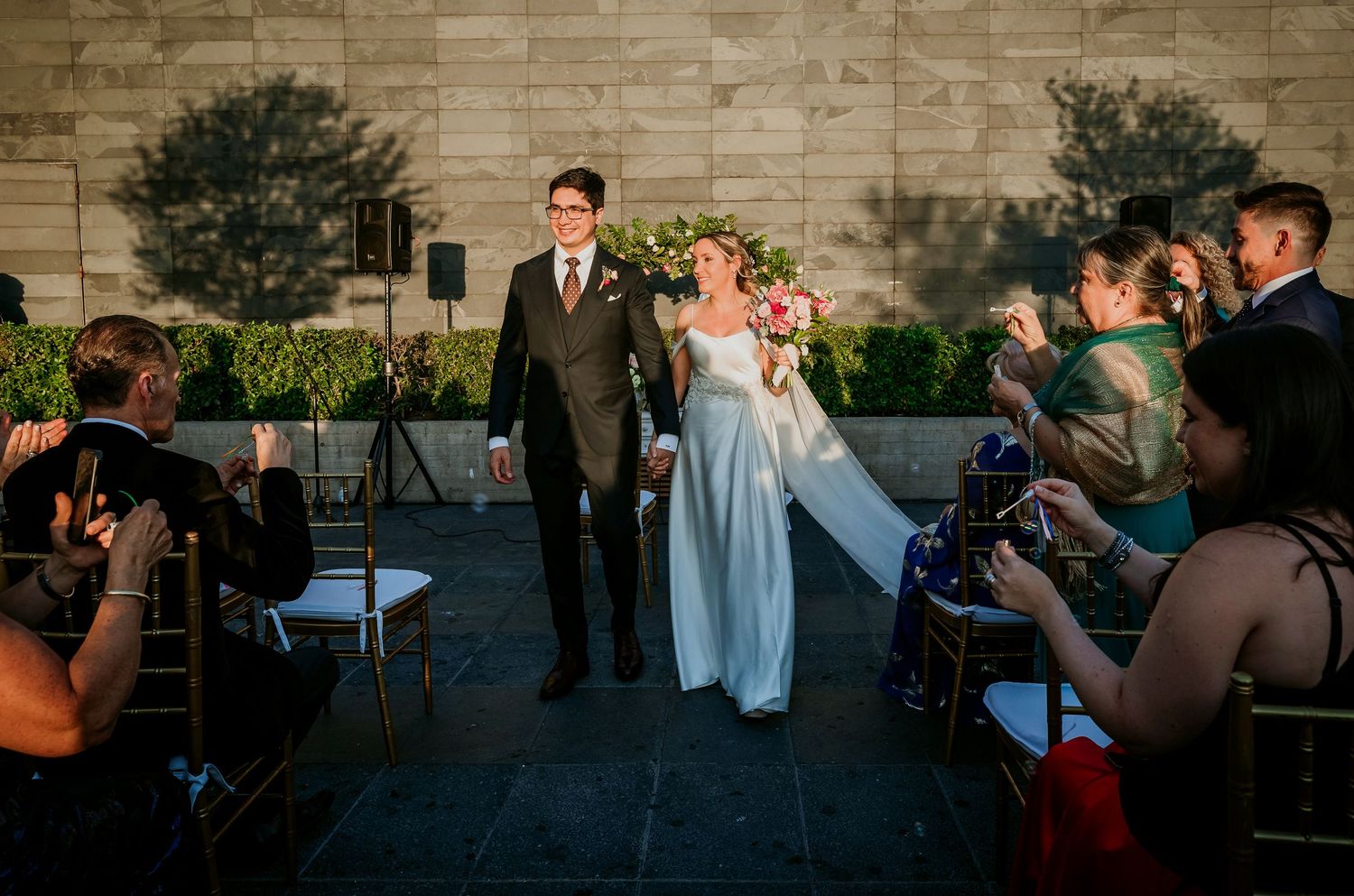 Newlyweds walk down aisle as guests celebrate at elegant outdoor wedding ceremony.
