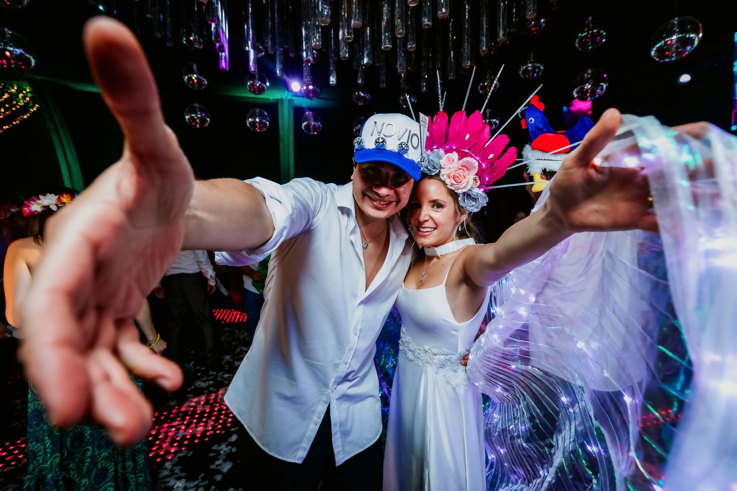 Smiling bride and groom reaching toward camera wearing festive headpieces at their wedding reception.
