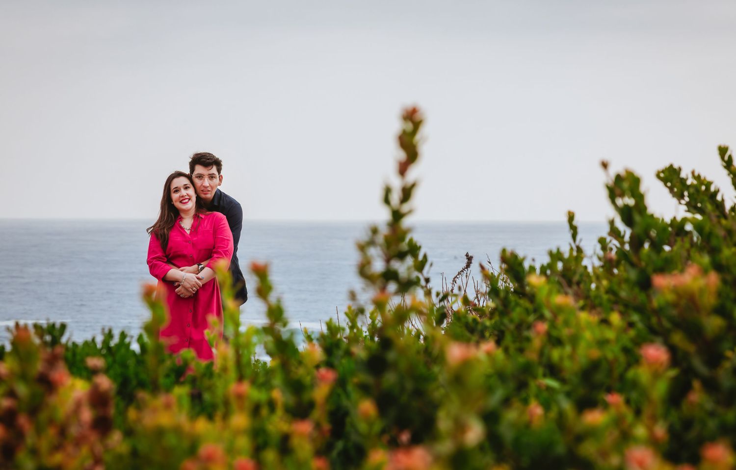 Couple embracing among colorful coastal shrubs with ocean backdrop, woman in pink dress and man in dark outfit.
