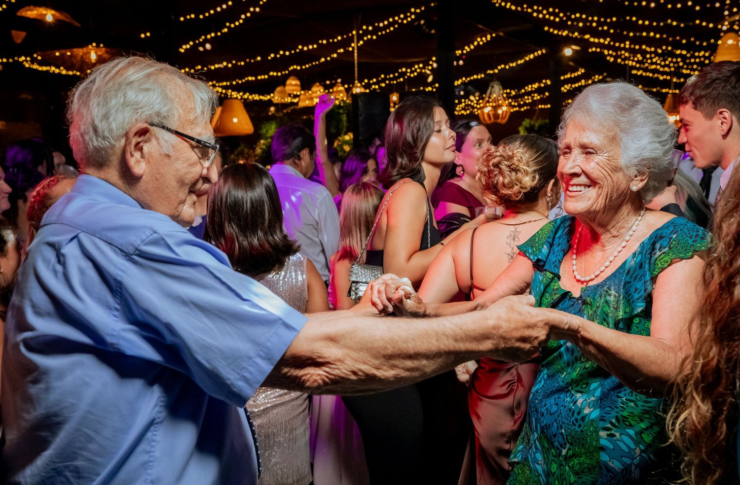 Elderly couple joyfully dancing together at vibrant wedding reception celebration with colorful lighting.