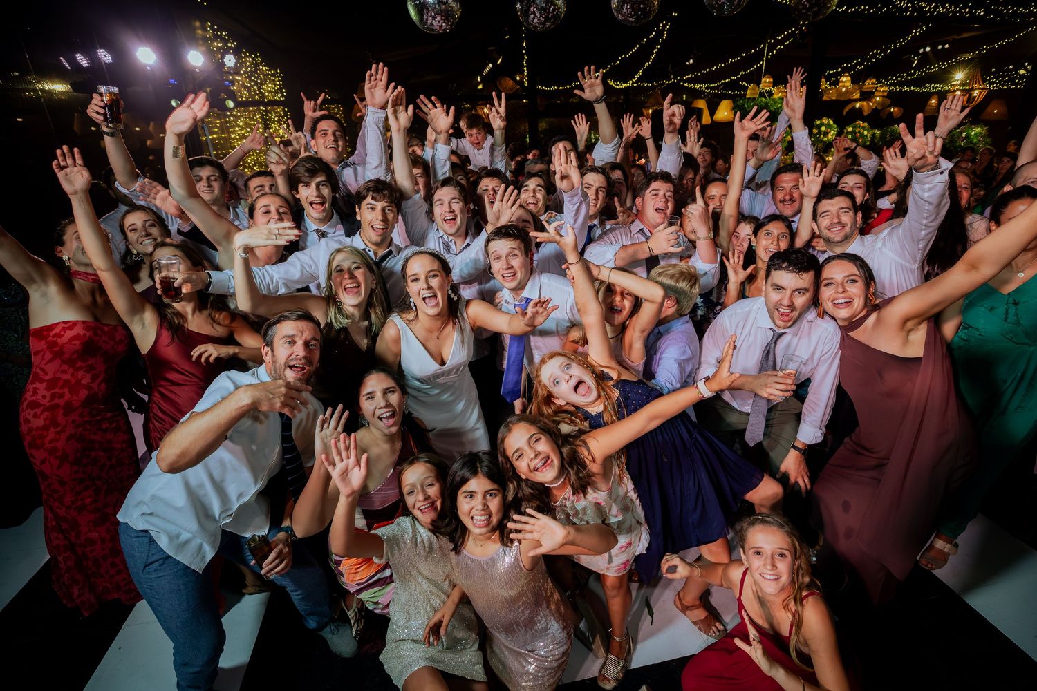 Large group of wedding guests raise hands and cheer together on dance floor.