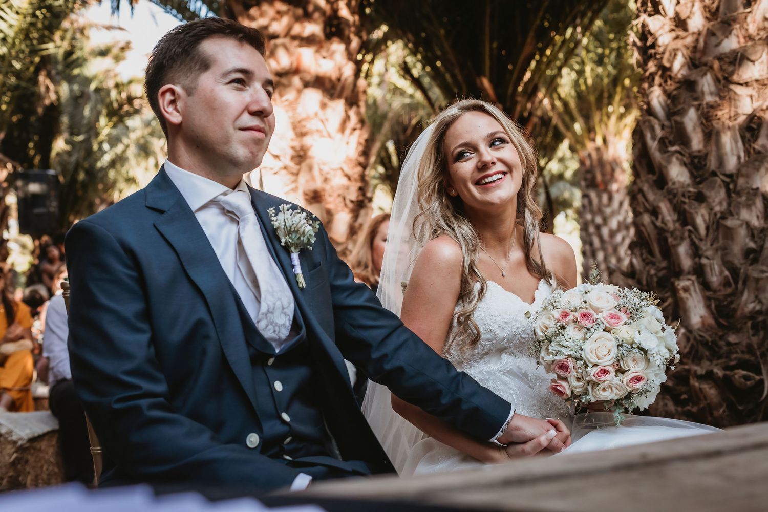 Bride in lace gown and groom in navy suit sit together smiling during outdoor wedding ceremony.