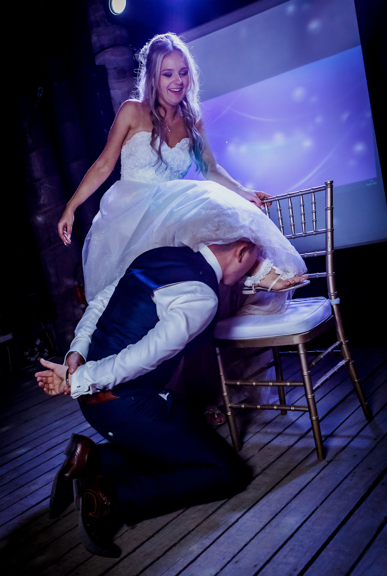 Bride seated on chair laughing as groom retrieves garter during fun wedding reception tradition.
