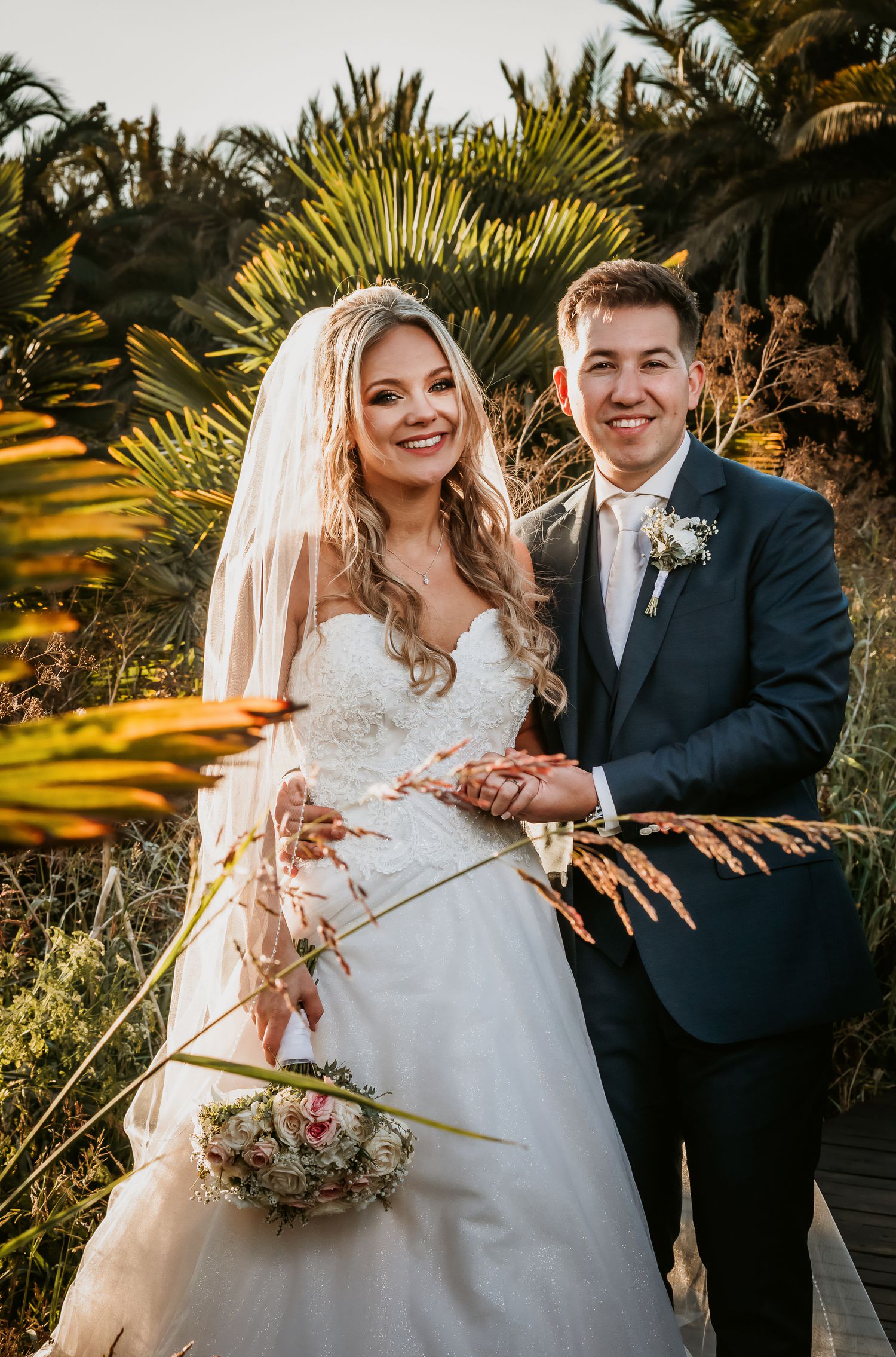 Bride and groom pose at golden hour among tropical palms, bride holding pastel bouquet.