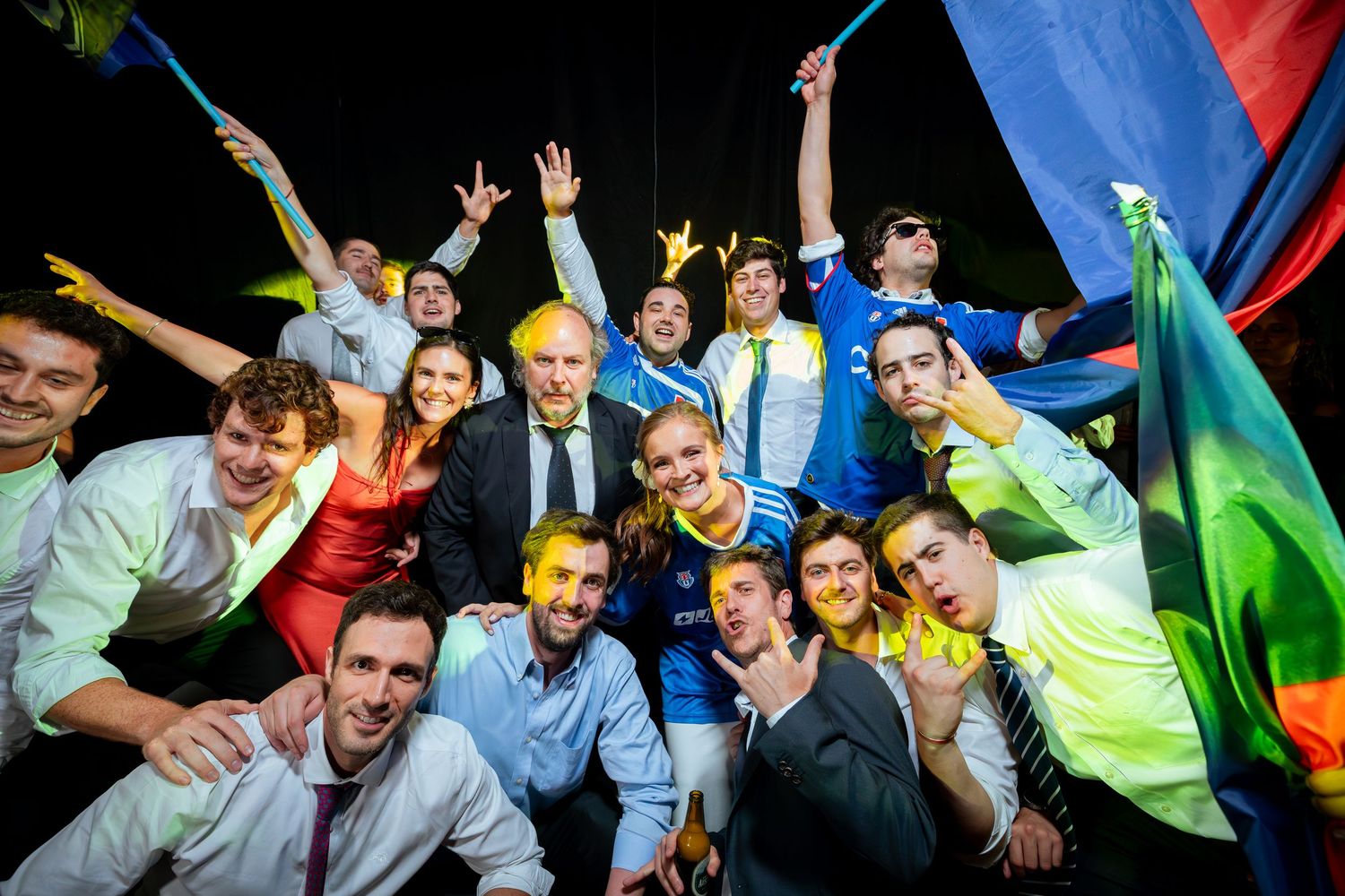 Large group of wedding guests posing joyfully with colorful flags at night celebration.