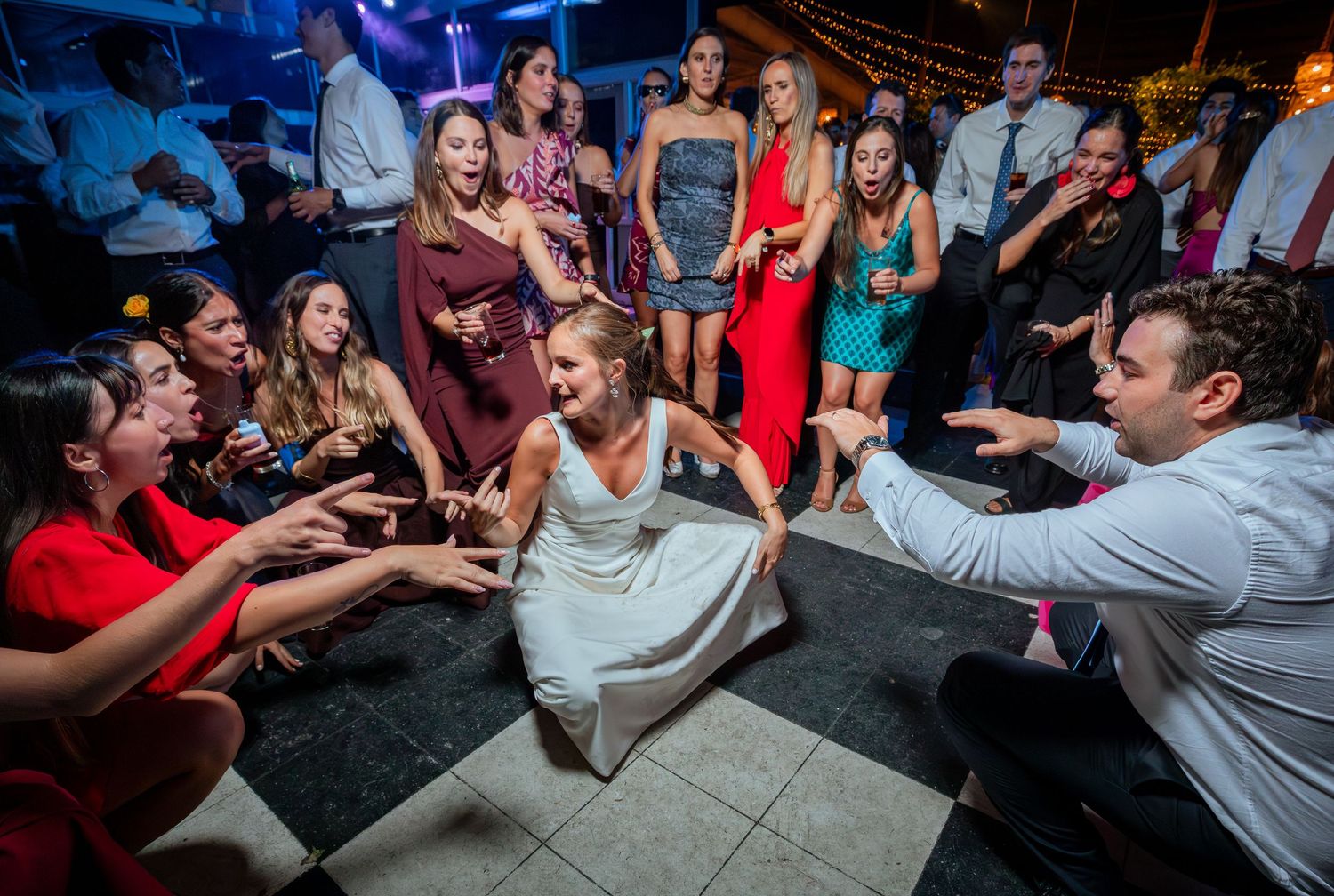 Bride in white dress dances joyfully on dance floor while wedding guests cheer and reach out around her.