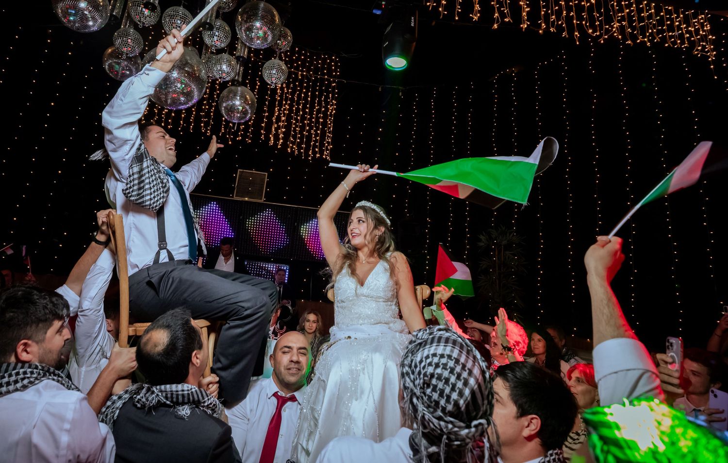 Bride and groom lifted on chairs during a lively wedding celebration with guests waving Palestinian flags.