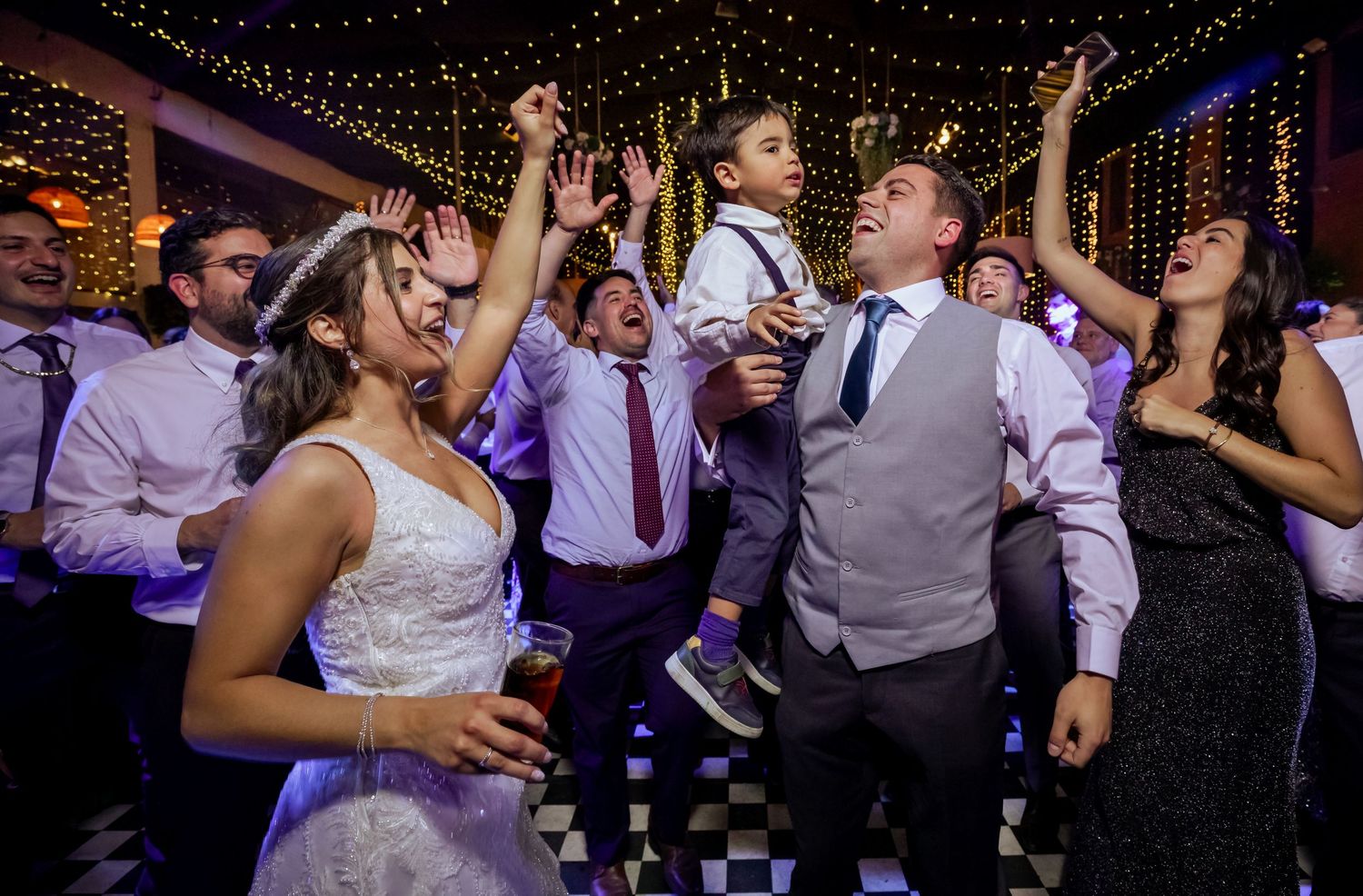 Bride and groom celebrate with guests on a festive wedding dance floor, a child joining the fun.