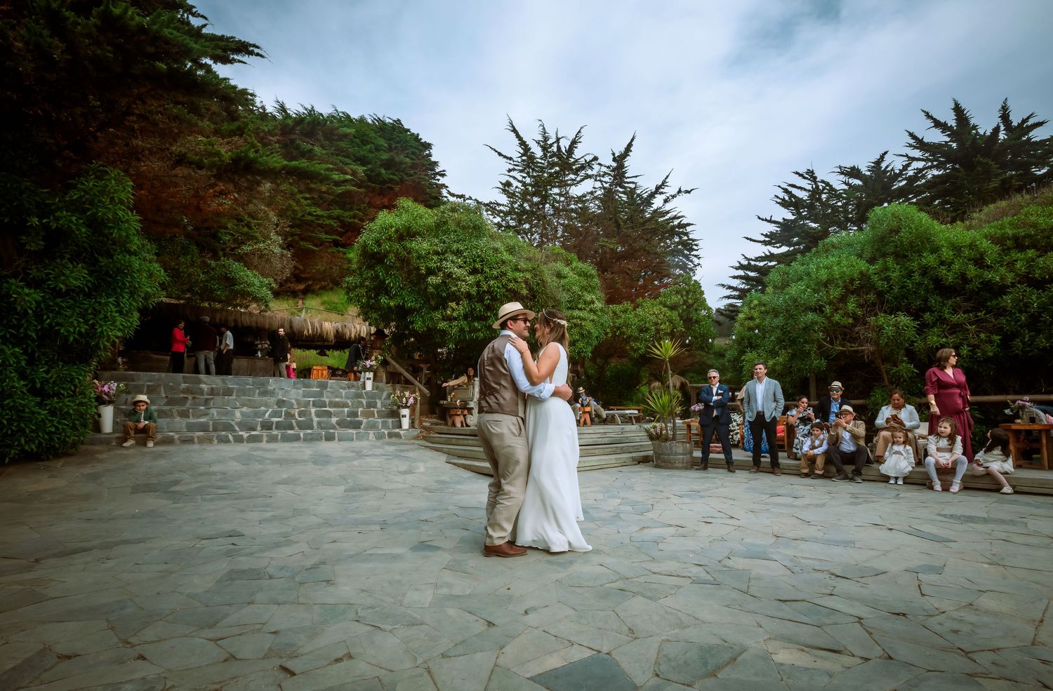 Bride and groom share their first dance on a stone patio as wedding guests watch in a scenic outdoor venue.