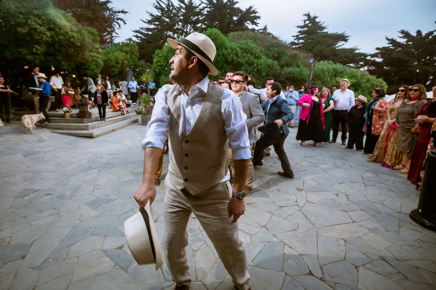 Groom in a white hat and vest walks confidently on a stone plaza during an outdoor wedding celebration.