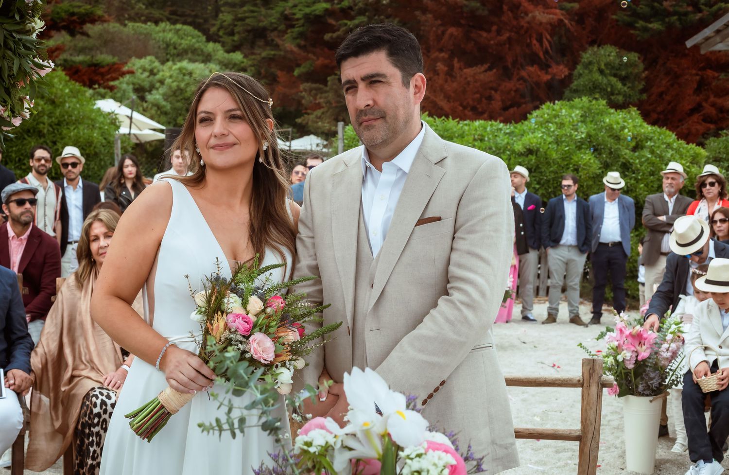 Smiling bride holding pink bouquet stands beside groom in beige suit at outdoor beach wedding.