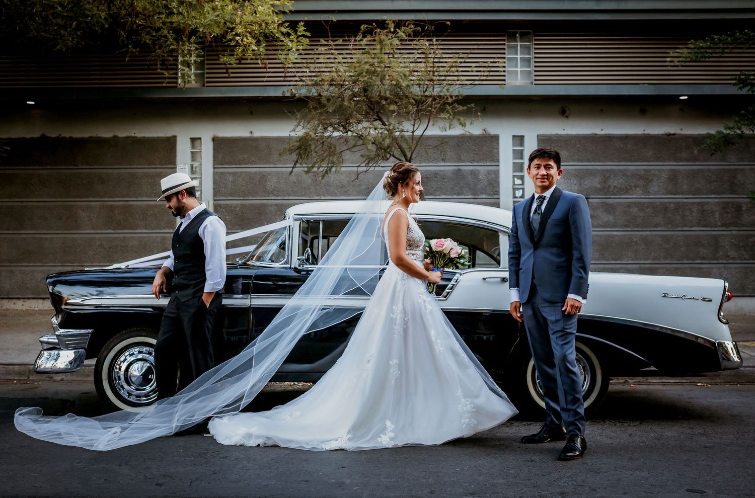 Bride and groom pose beside a classic vintage black and white car with a suited companion nearby.