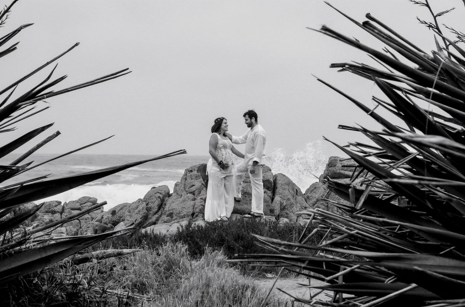 Black and white photo of a couple holding hands on a rocky hillside, framed by tropical plants.