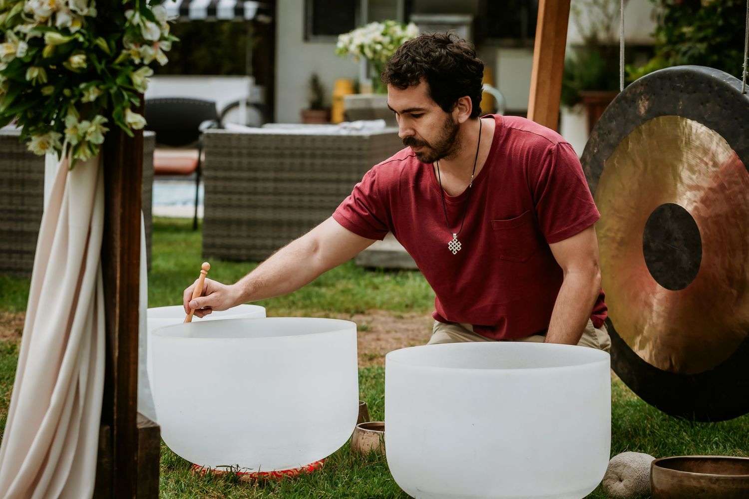 Man playing crystal singing bowls outdoors near a large gong during a sound healing session in a garden setting.