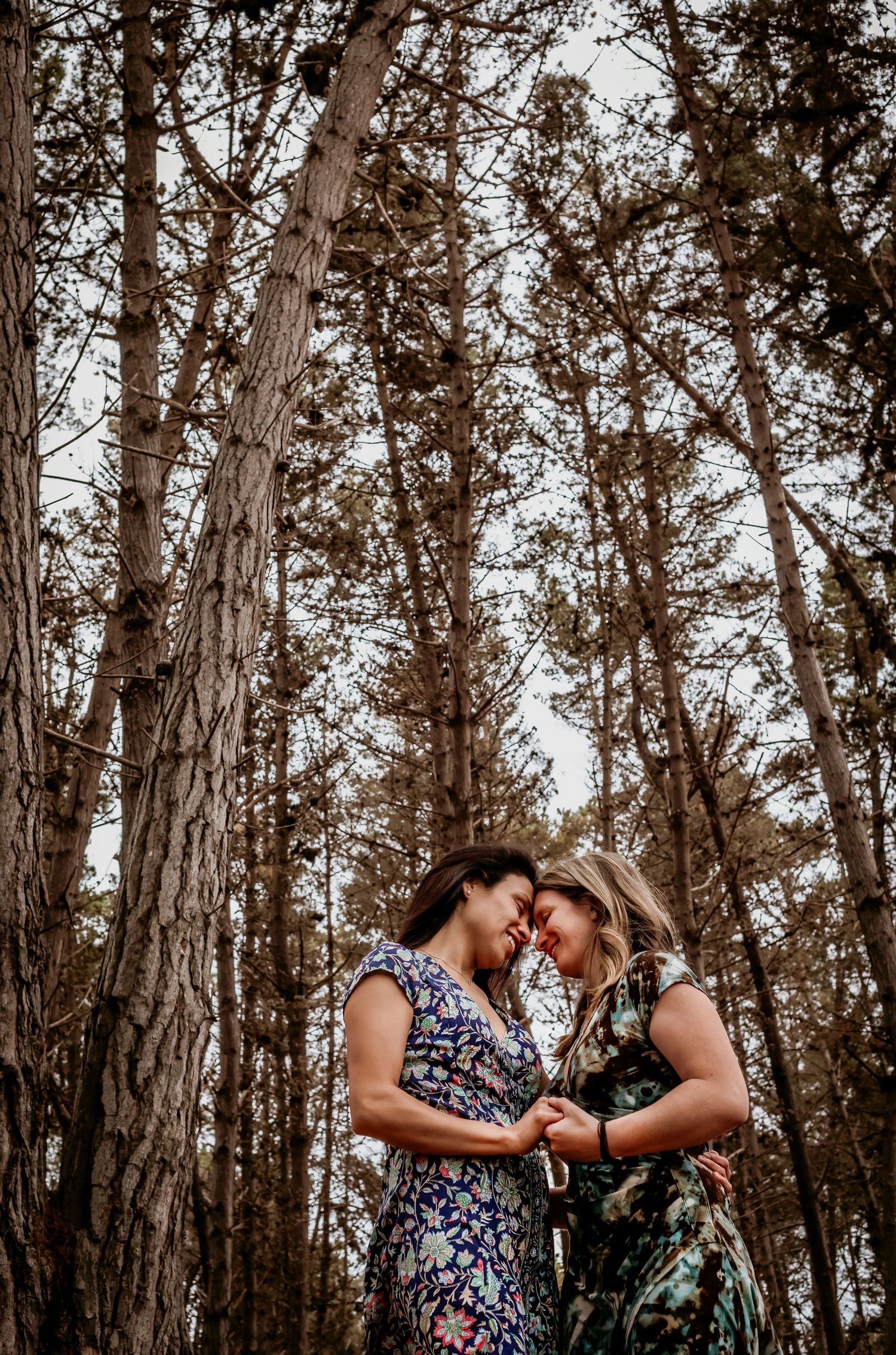 Two women share a romantic forehead touch while embracing beneath tall pine trees outdoors.