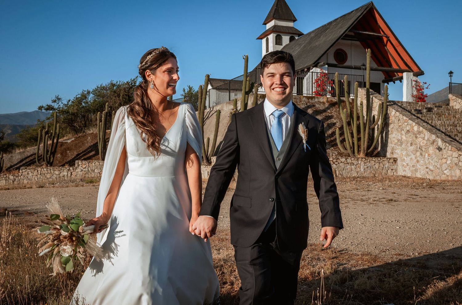 Smiling bride and groom walking hand in hand near charming rustic chapel with cacti.