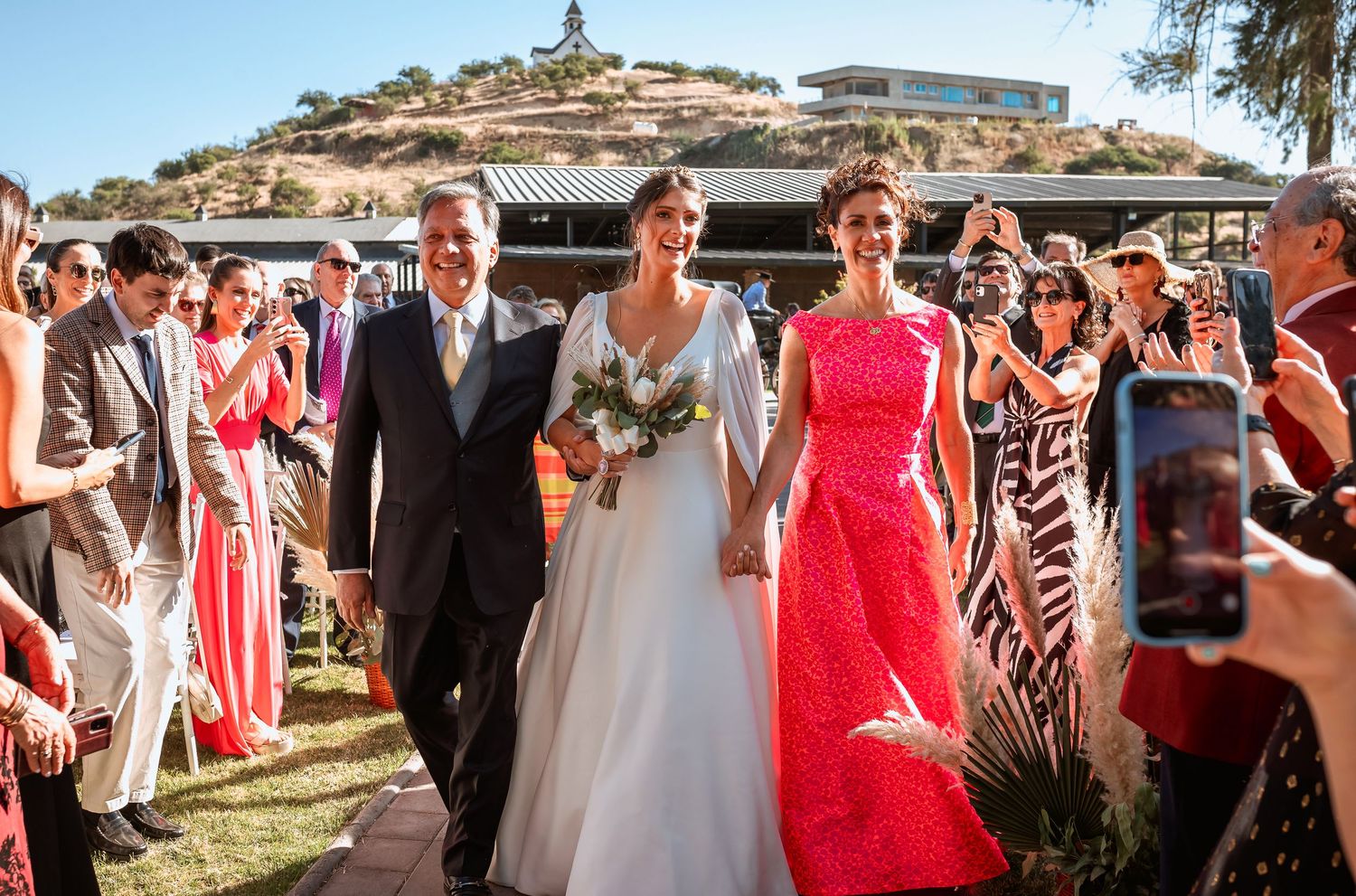 Bride walks down outdoor aisle escorted by parents at sunny wedding.