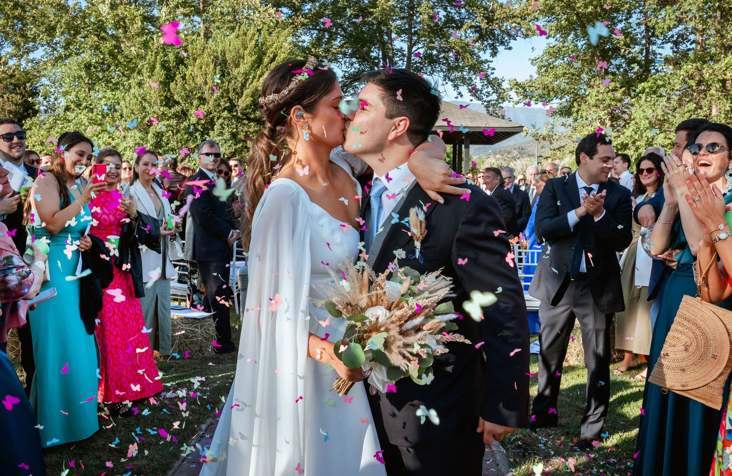 Newlyweds sharing romantic kiss surrounded by colorful confetti and cheering wedding guests outdoors.