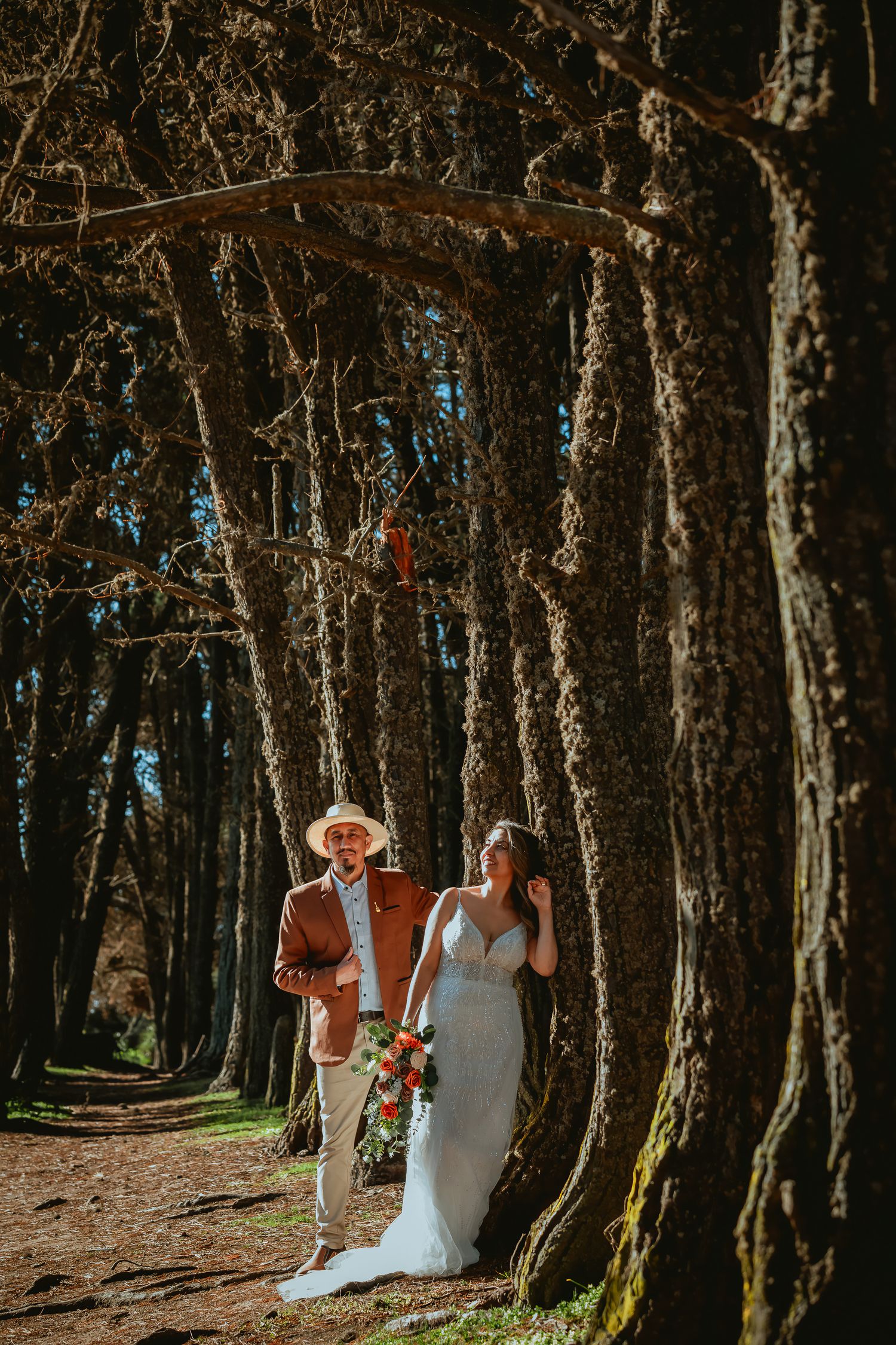Bride in a white gown holding an orange bouquet walks with a groom in a rust blazer and straw hat among tall forest trees.