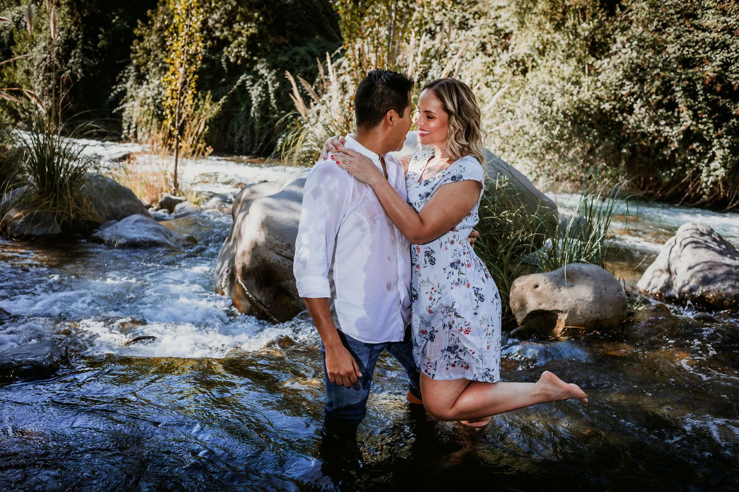 Couple embracing romantically in a shallow rocky stream surrounded by lush green trees and boulders outdoors.