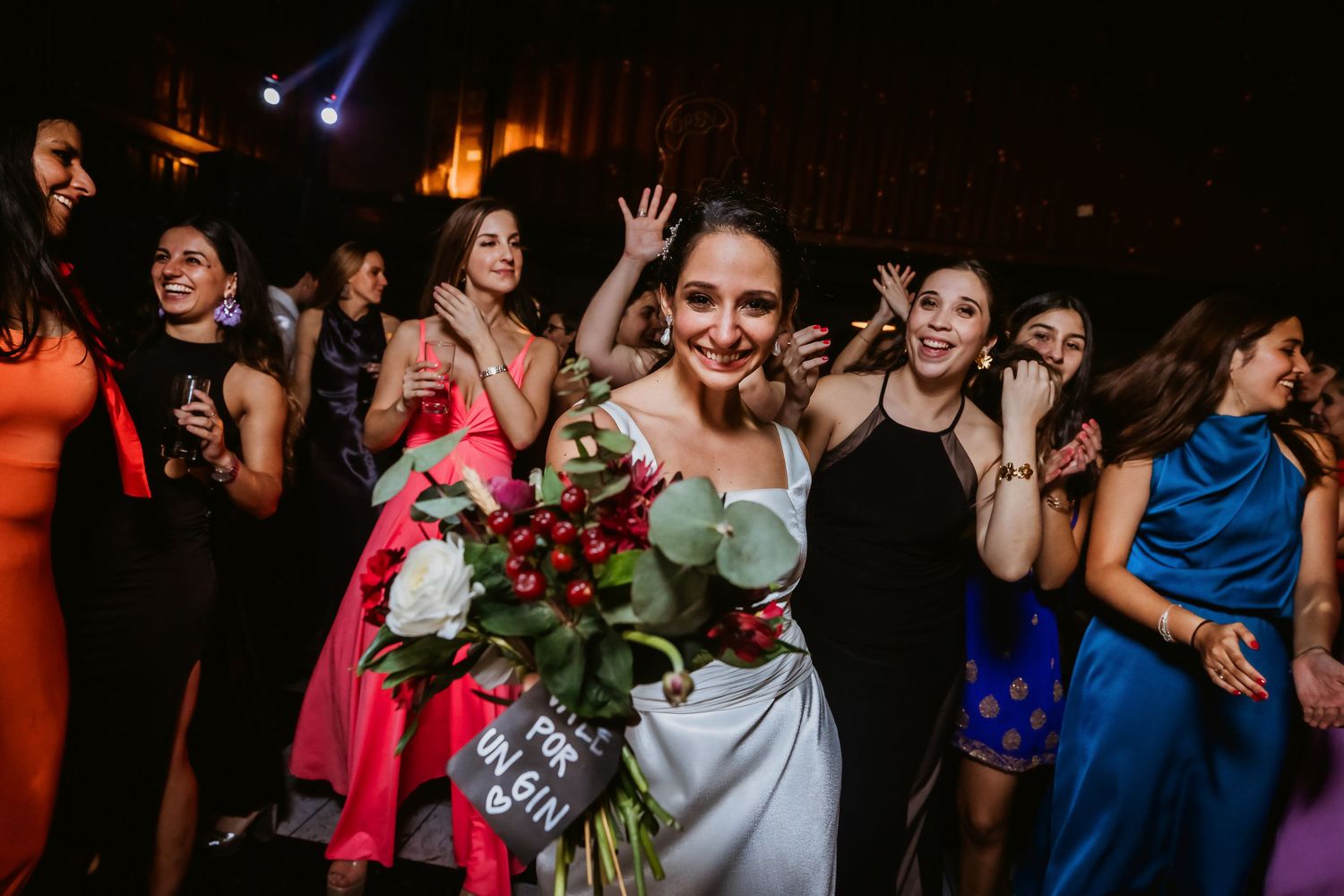 Bride holding red and white bouquet dancing with guests in colorful dresses at a lively wedding reception.