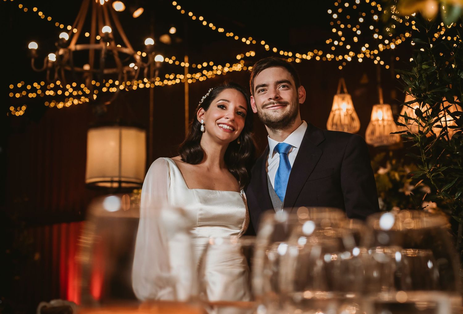 A smiling bride and groom pose together at their wedding reception under warm string lights and lanterns.