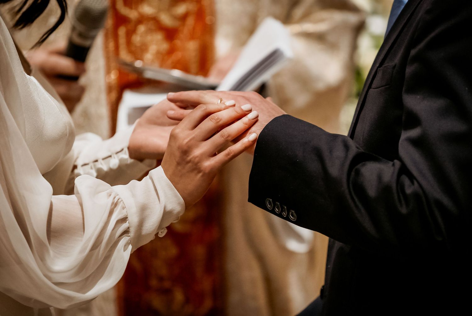 Groom placing a wedding ring on bride's finger during a ceremony, with officiant holding book in background.