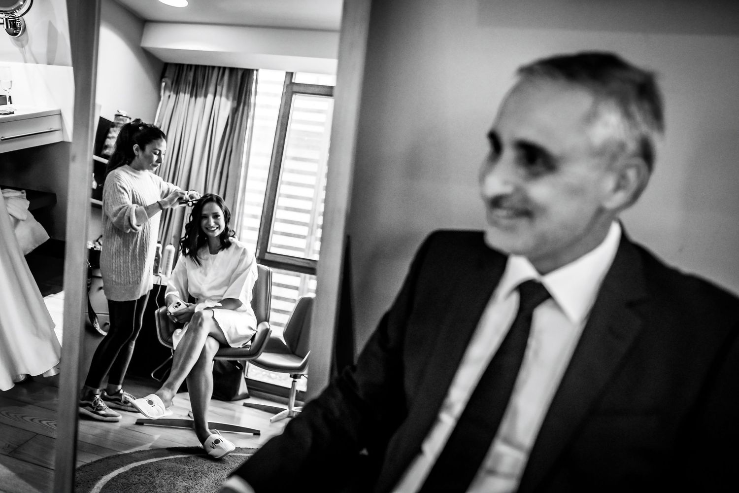 Father of the bride watches as hairstylist prepares bride on her wedding day in black and white photo.