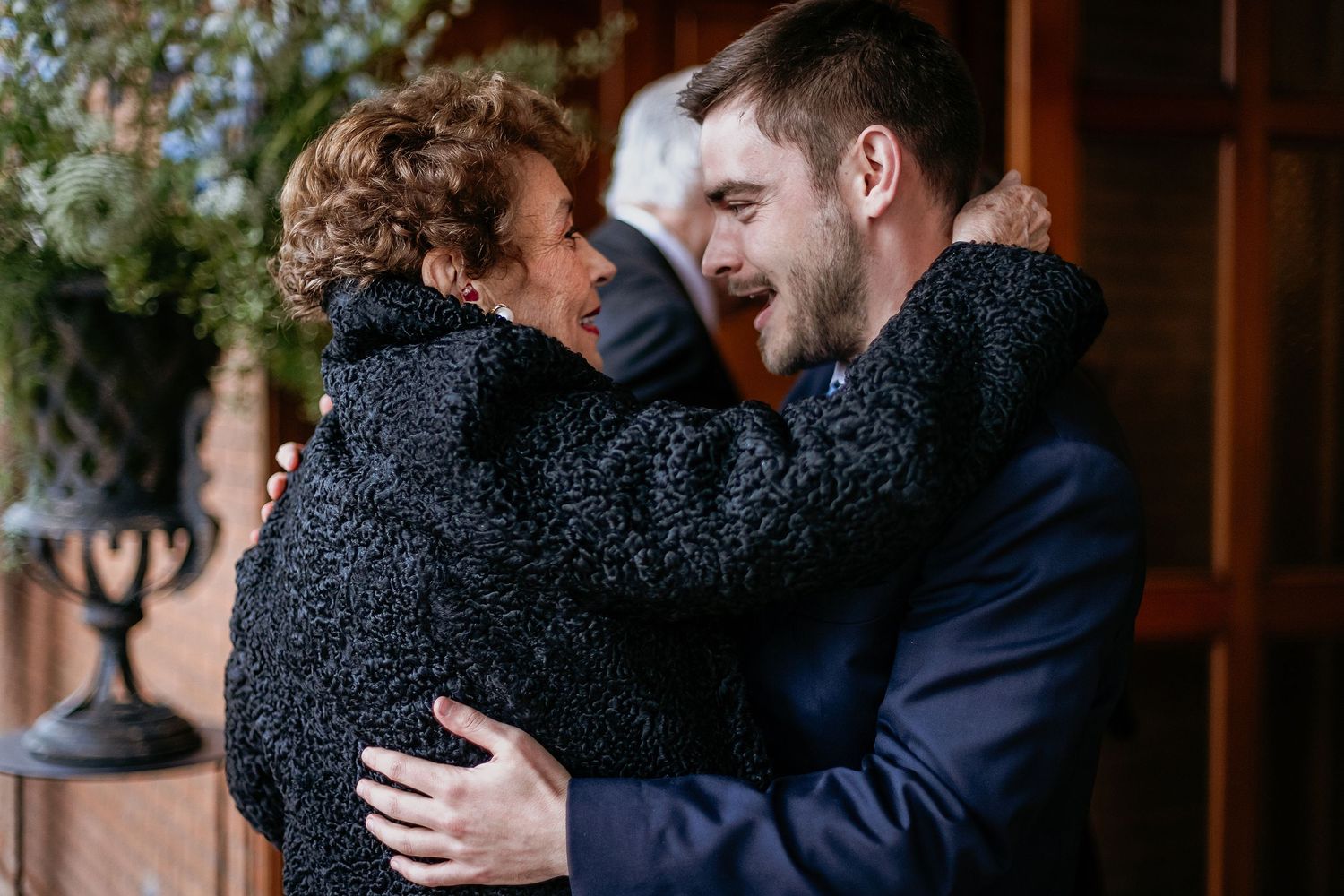 Two people sharing a warm embrace outdoors, smiling joyfully, dressed in elegant attire at a special occasion.