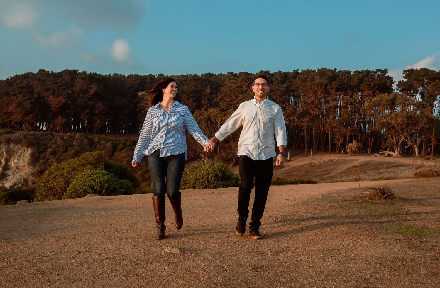 Couple holding hands walking on a scenic coastal path with trees and blue sky.