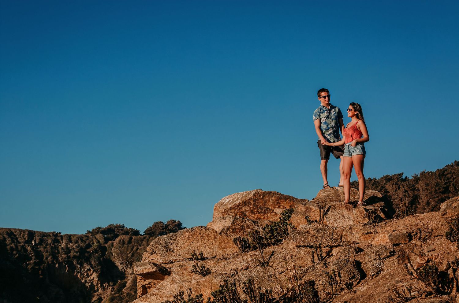 Couple standing together on rocky terrain against clear blue sky.
