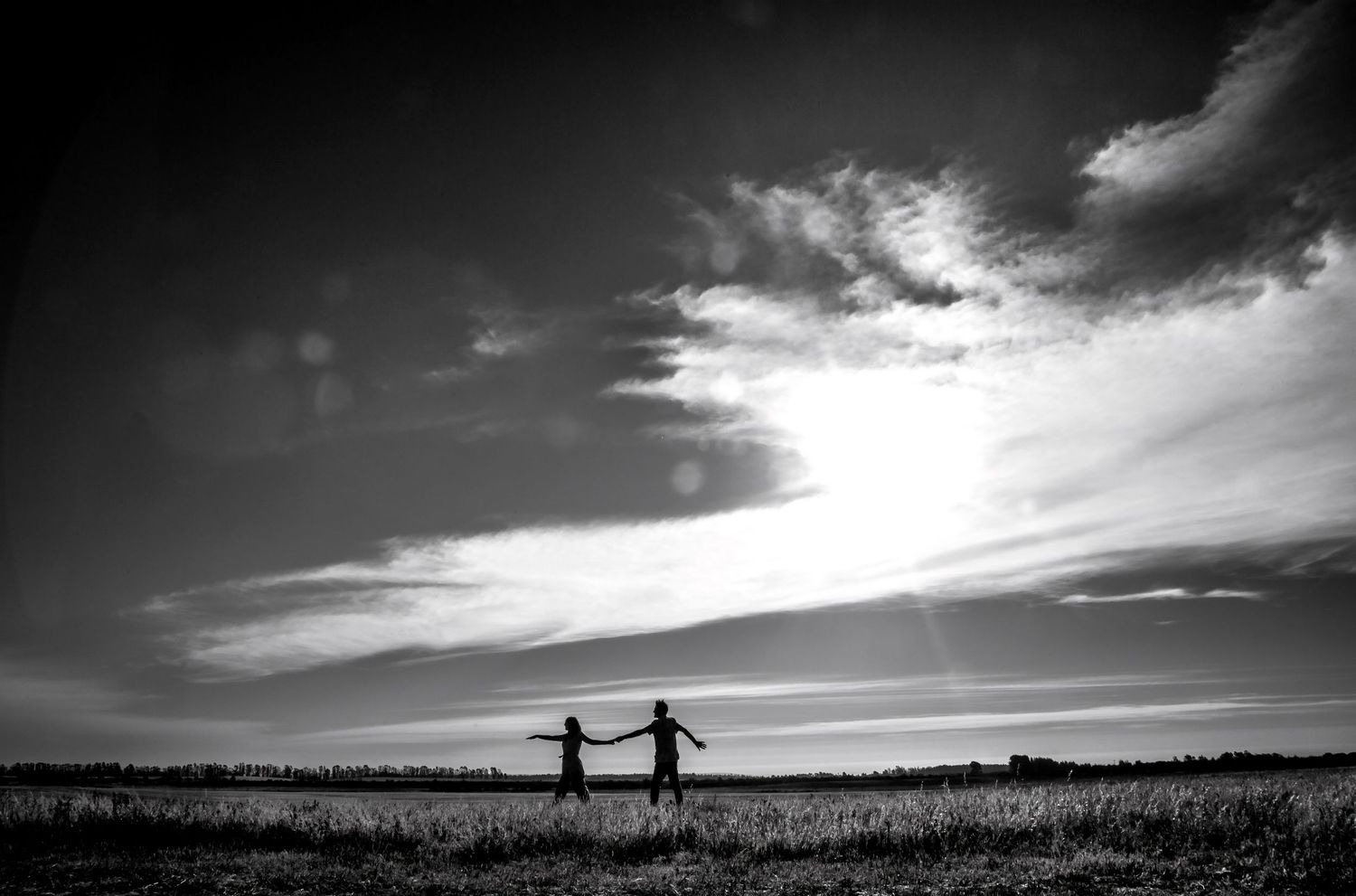 Black and white silhouette of a couple holding hands in an open field under a vast sky.