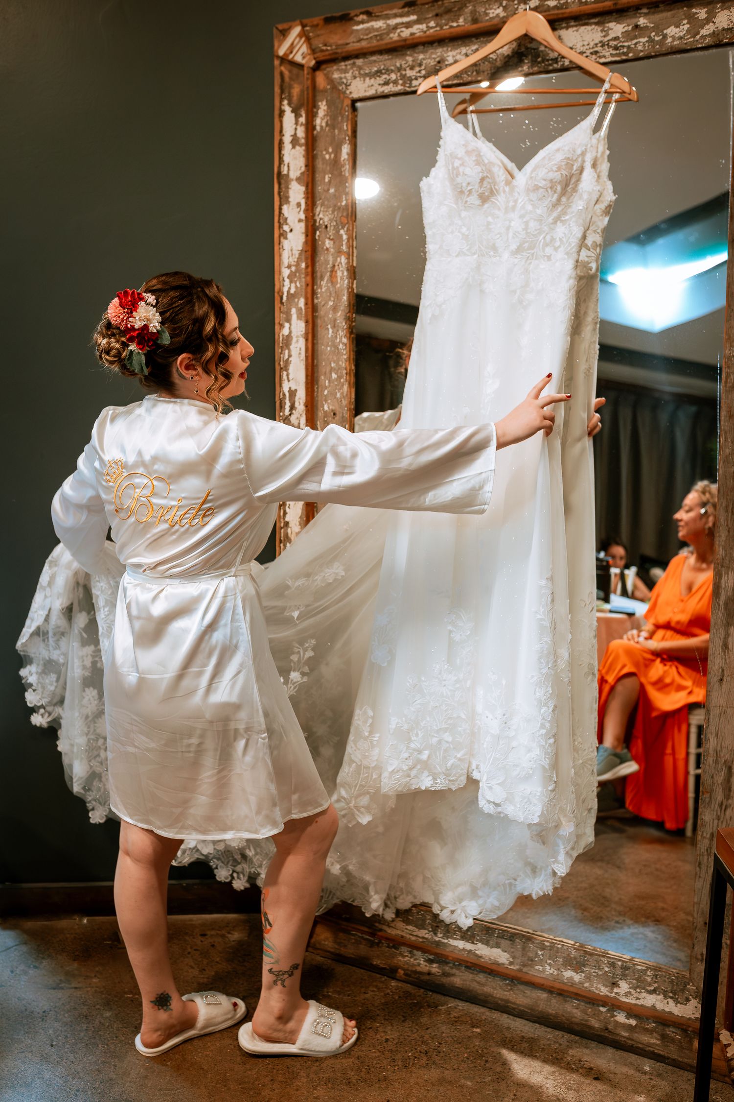 Bride in white satin robe reaching for lace wedding gown hanging on ornate mirror frame.