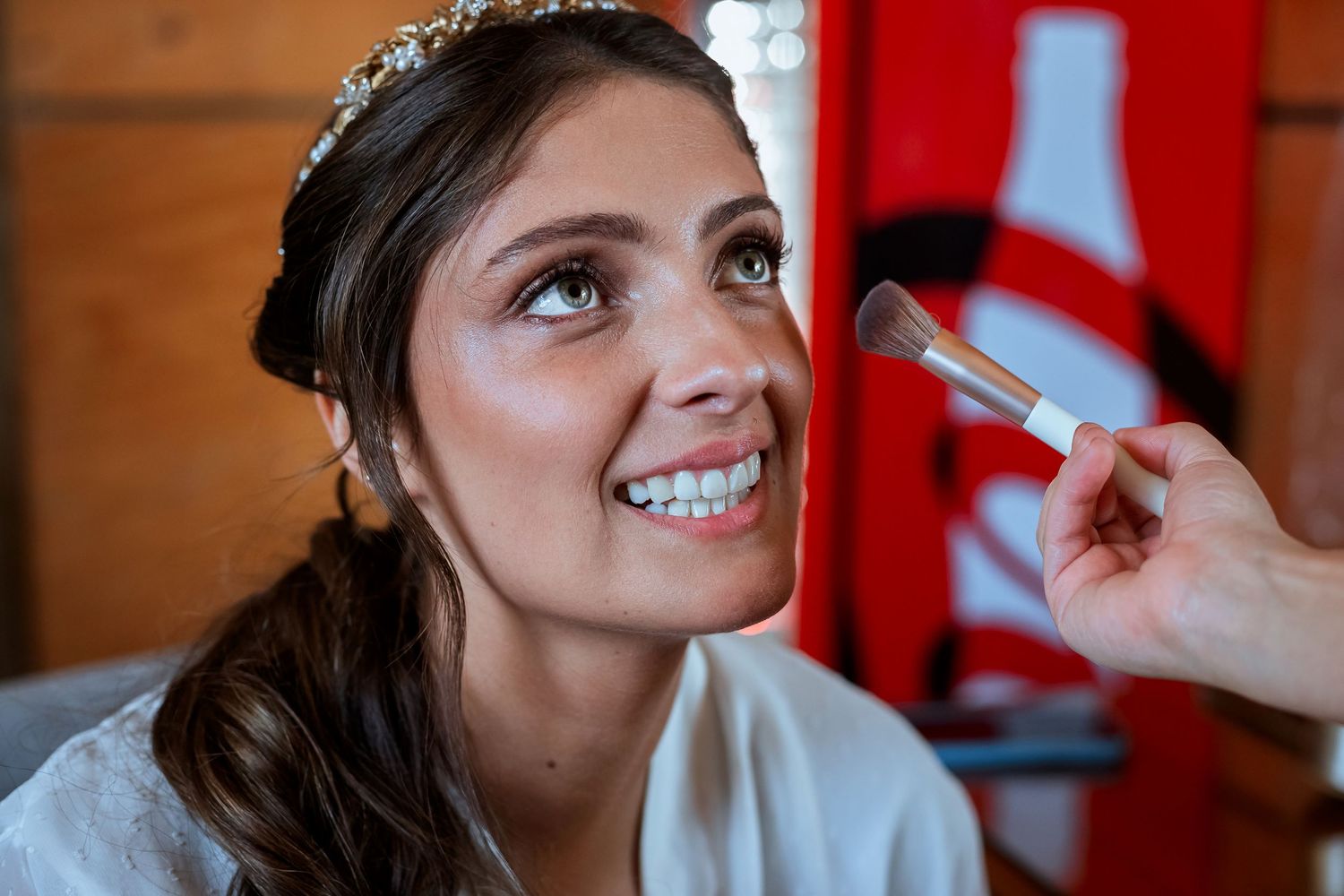 Smiling bride wearing a tiara getting makeup applied with a brush during wedding day preparations.