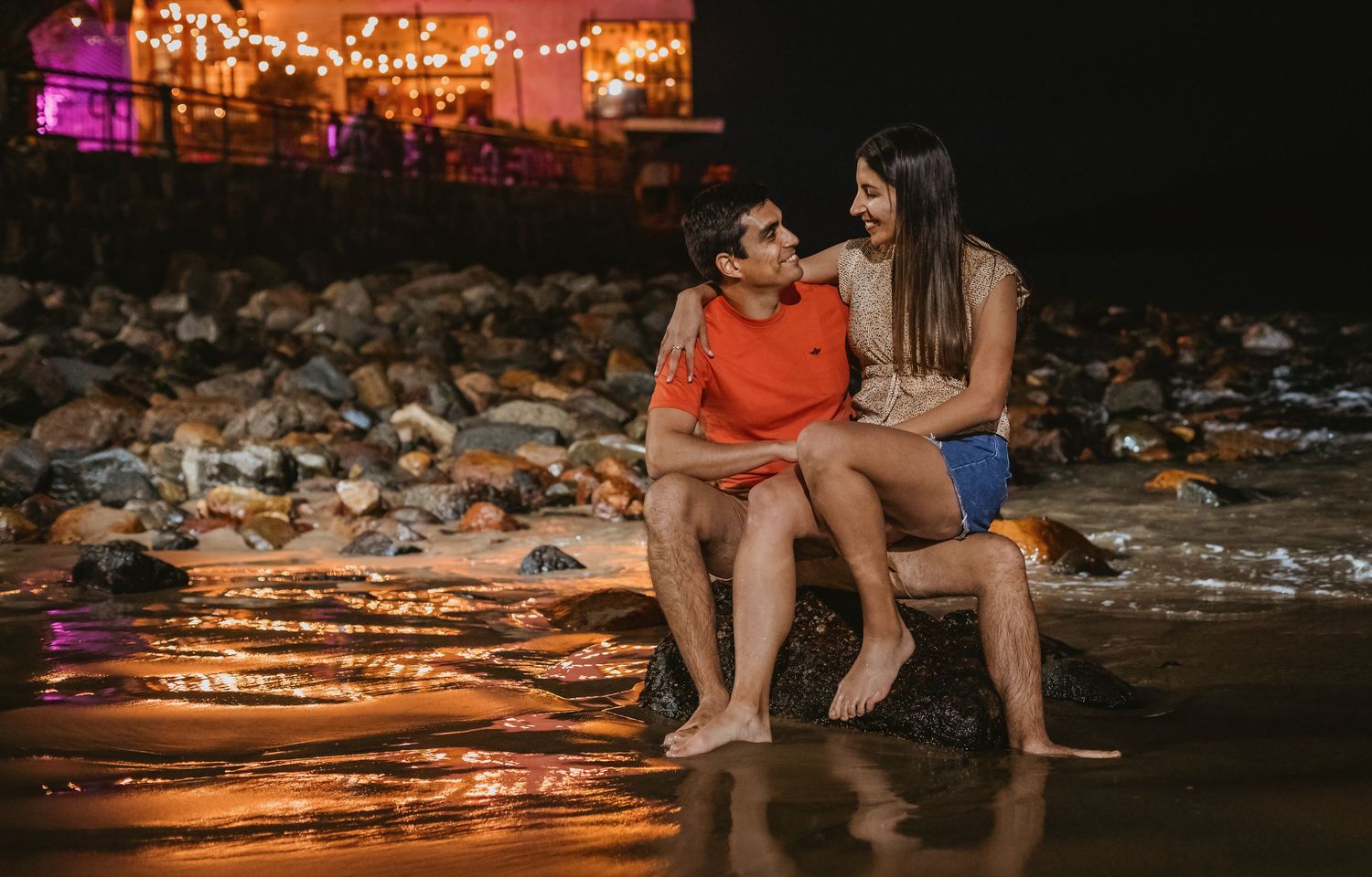 Couple sitting on rocks at night near a beach with warm string lights glowing behind them.