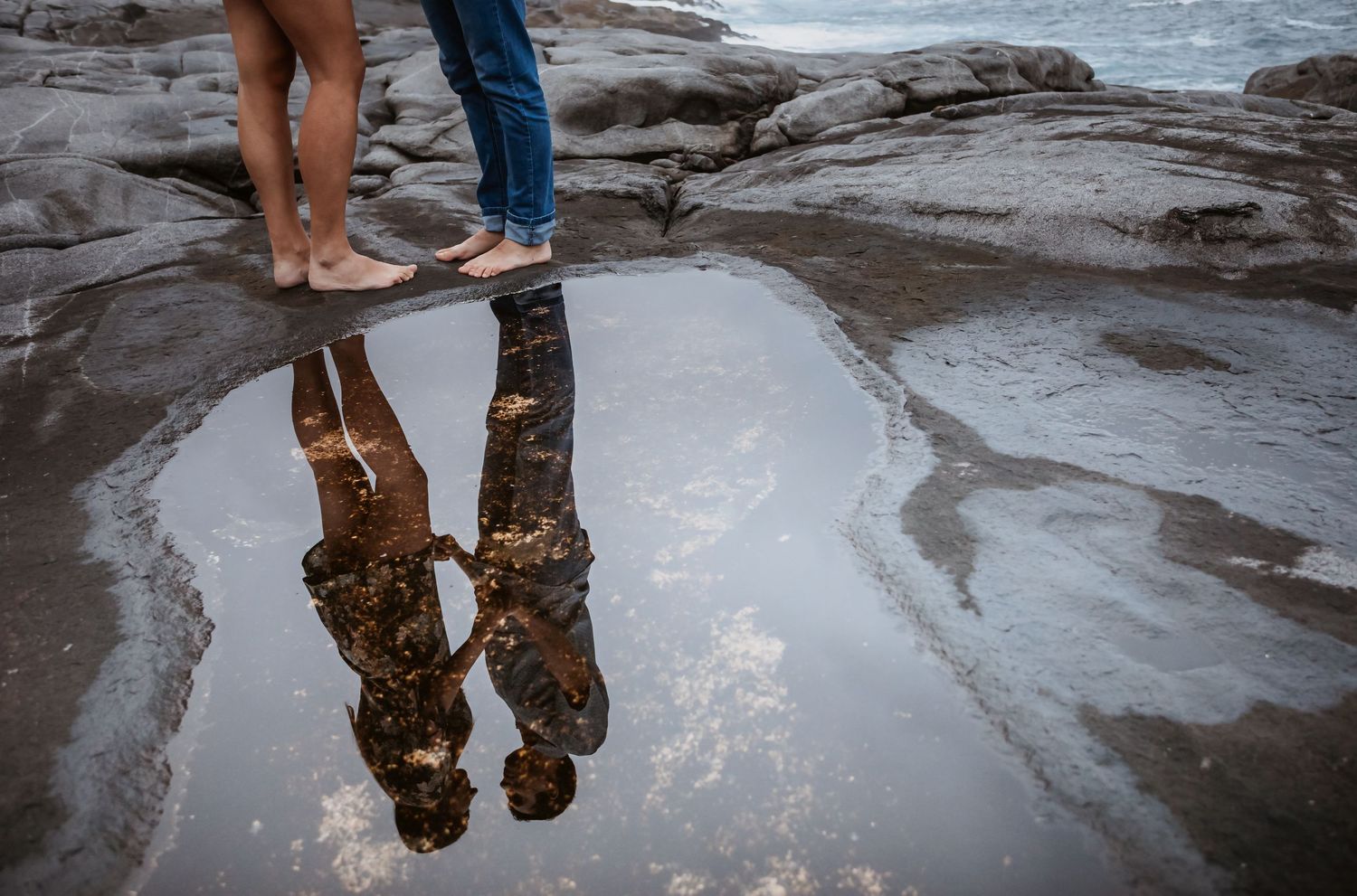Reflection of a couple standing on wet rocks captured in a natural tide pool puddle.