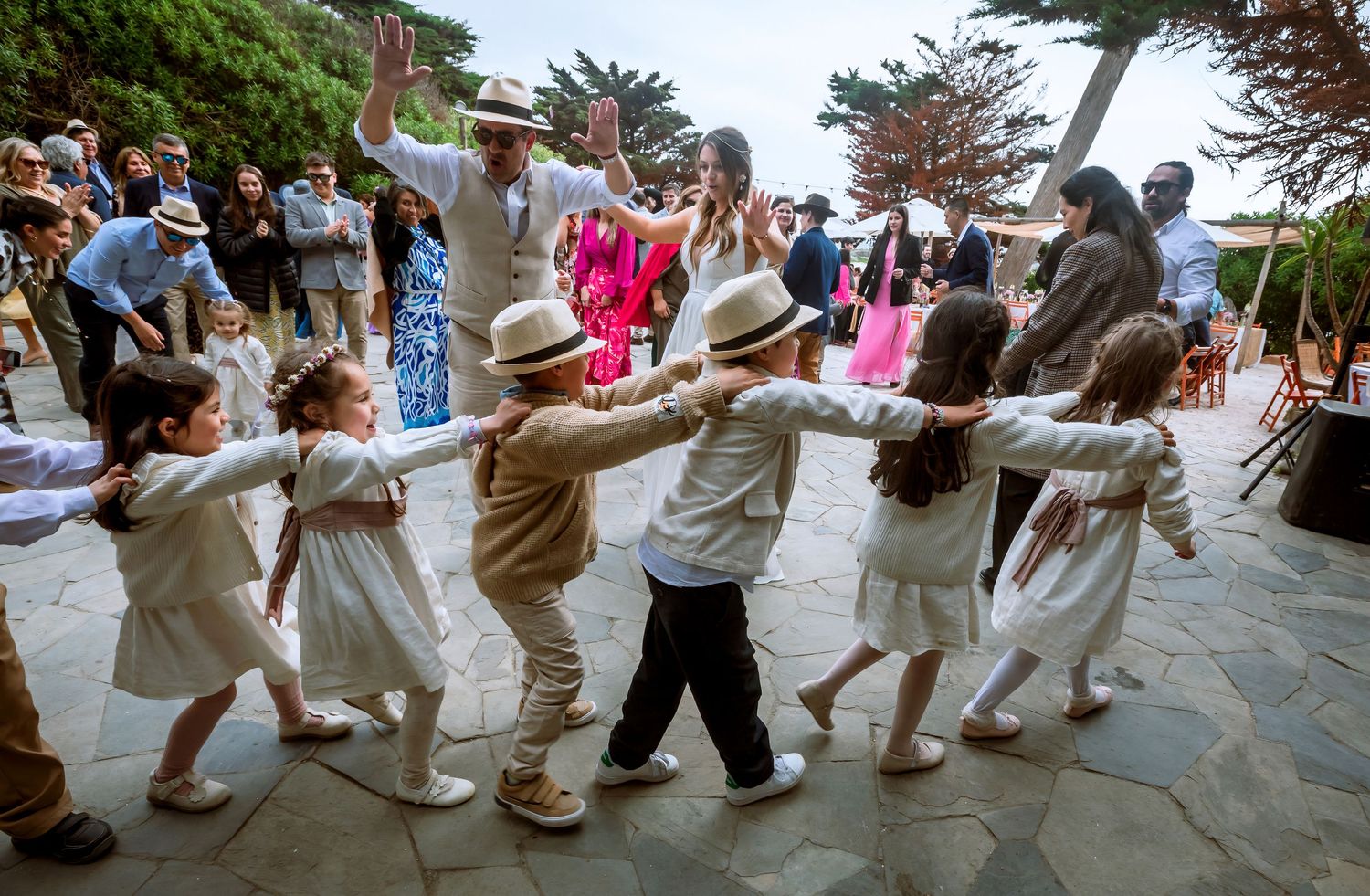 Kids holding hands in a circle dance at an outdoor wedding celebration with adults watching.