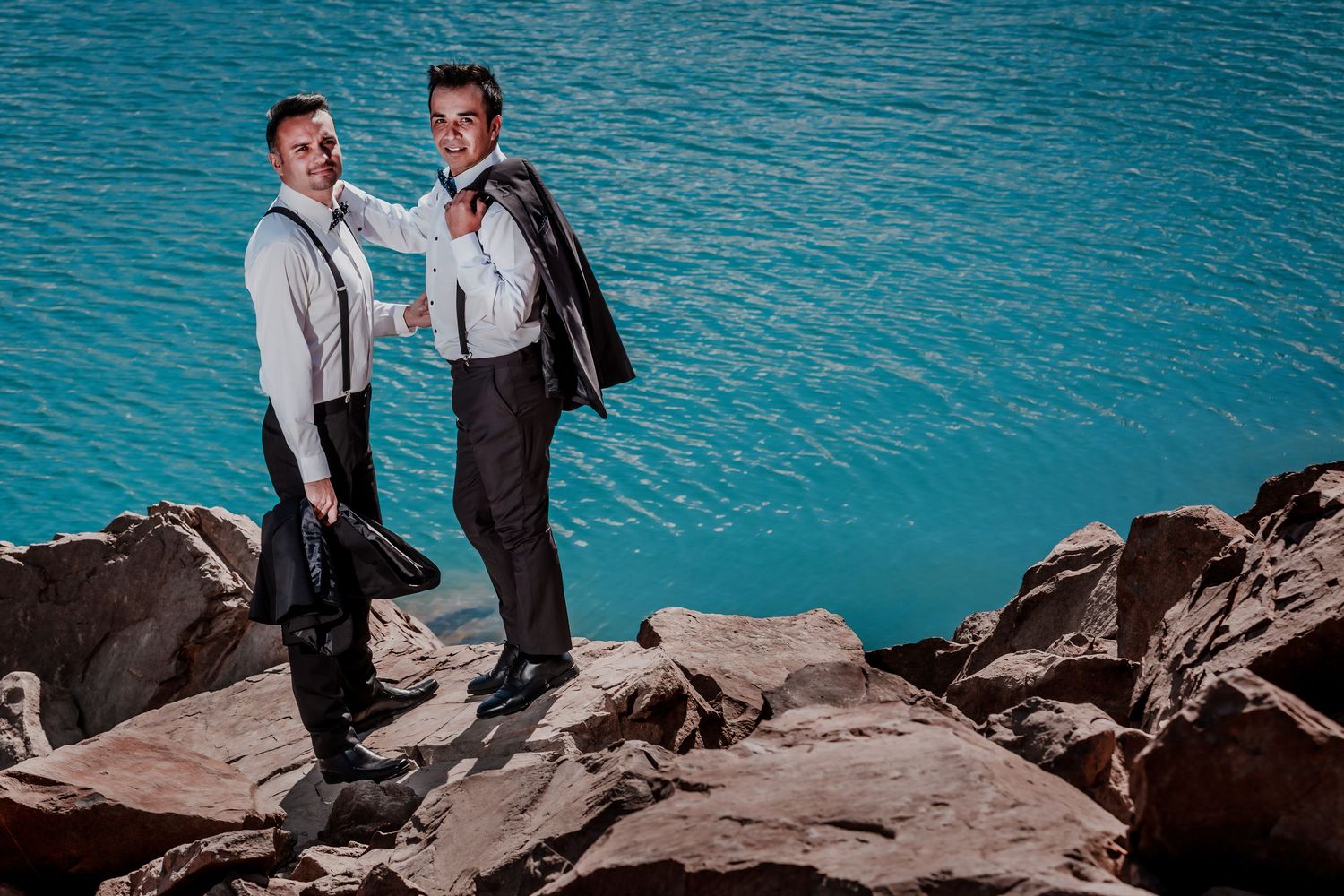 Two grooms in formal suits stand on rocky shore beside a vibrant turquoise mountain lake.