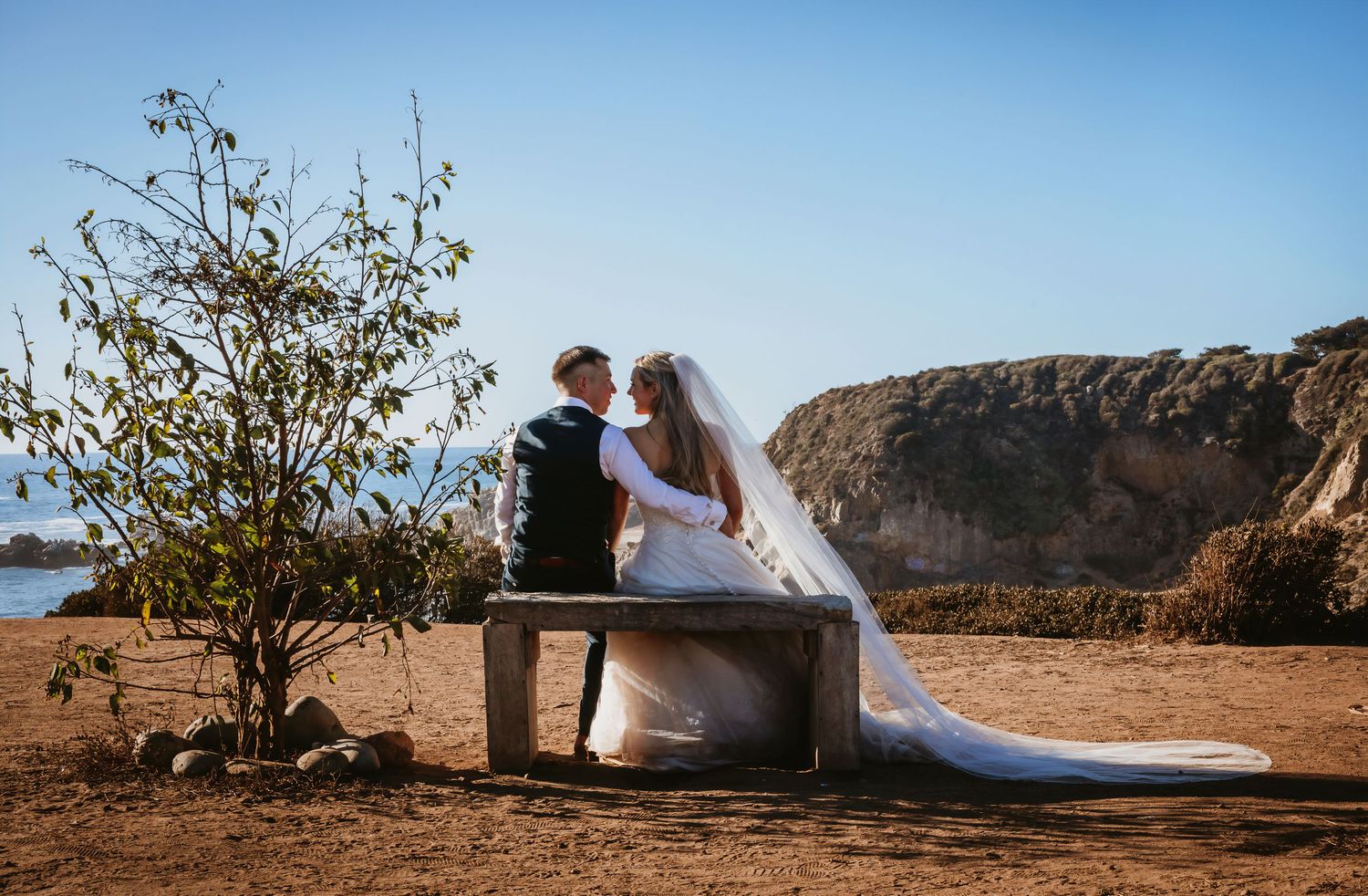 Bride and groom sitting on a wooden bench sharing a kiss overlooking the ocean on their wedding day.