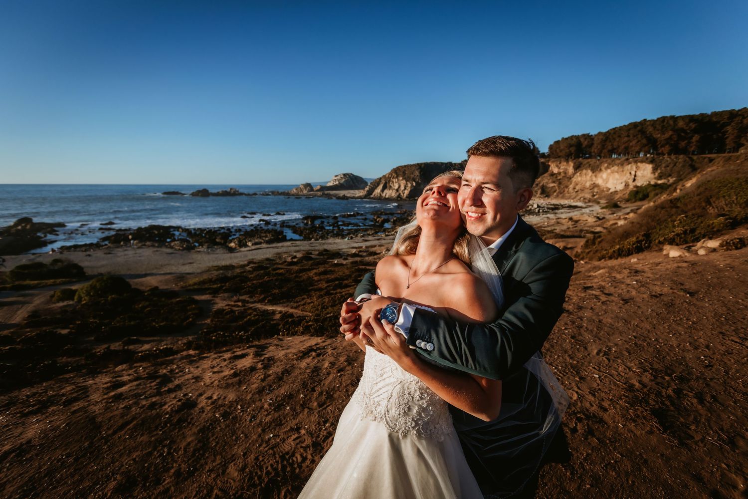 Couple embraces warmly on scenic coastal bluff with rocky beach and ocean cliffs in background.