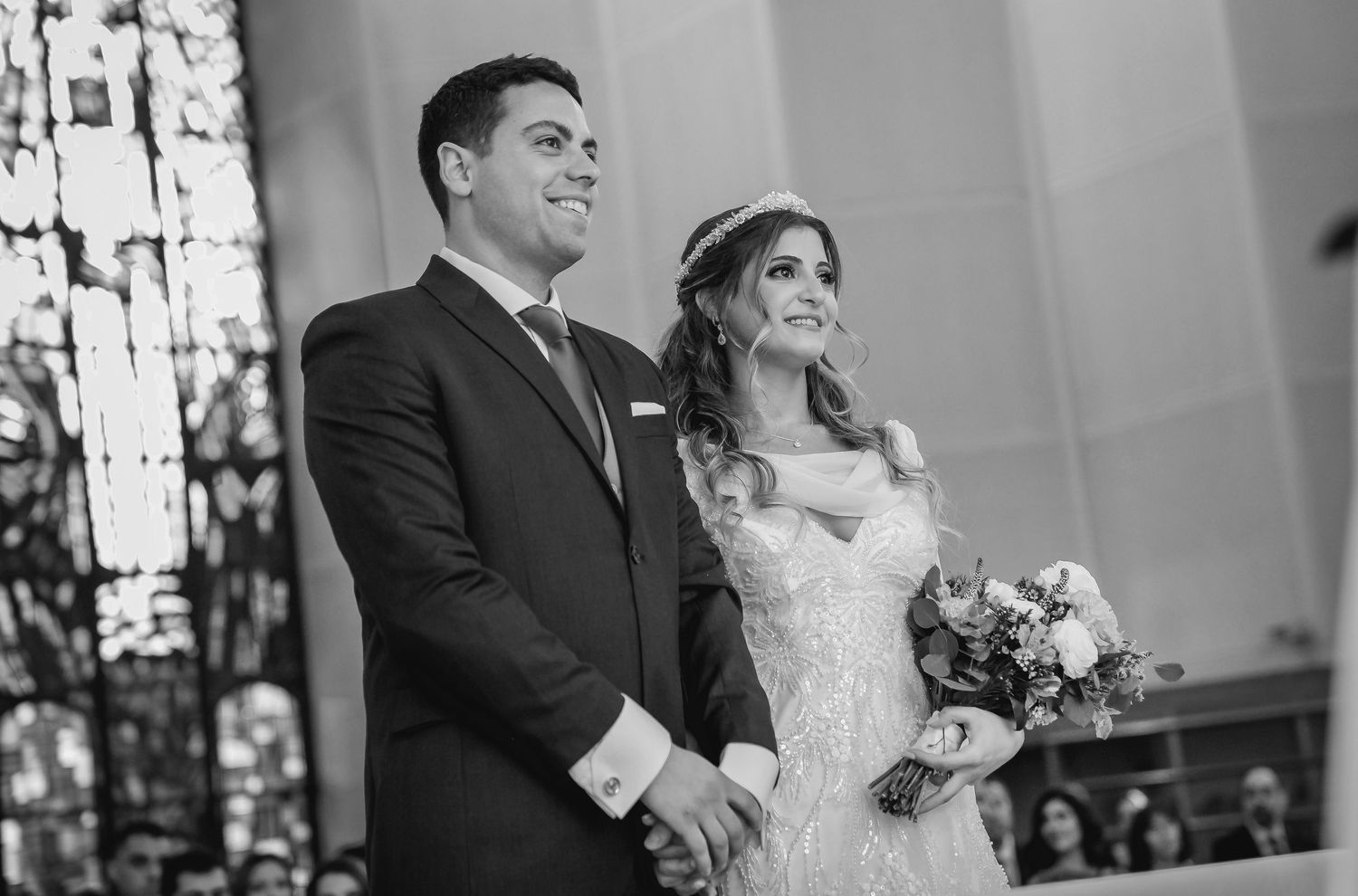 Black and white photo of smiling bride holding bouquet and groom standing together at church altar.