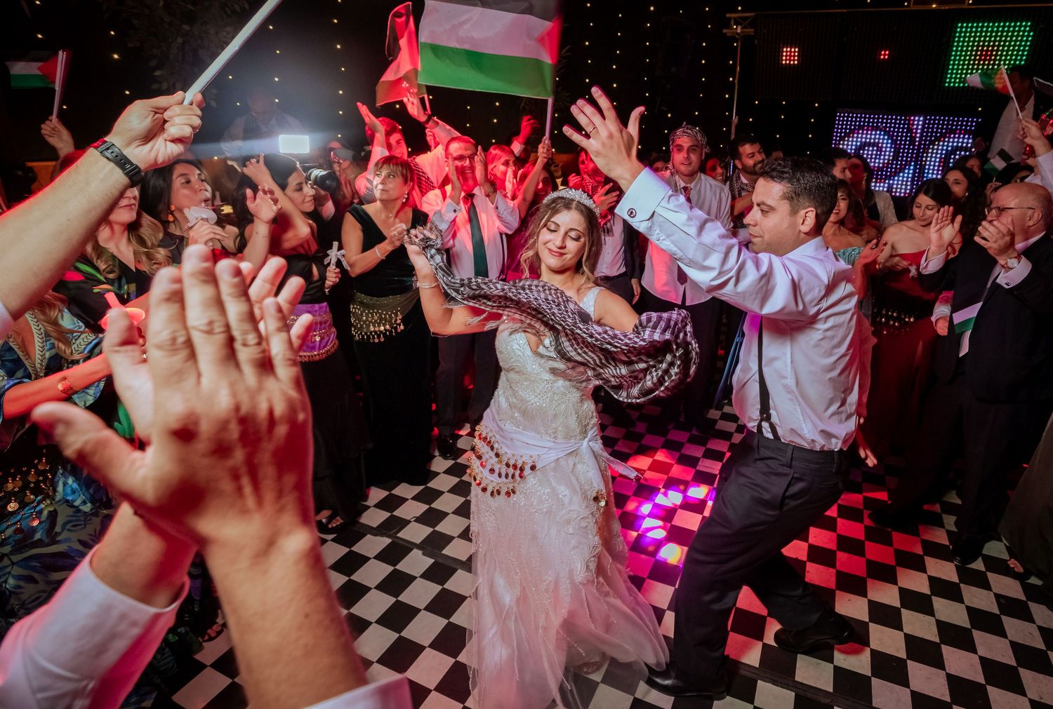 Bride and groom dance joyfully at a vibrant wedding reception with guests waving flags and celebrating.