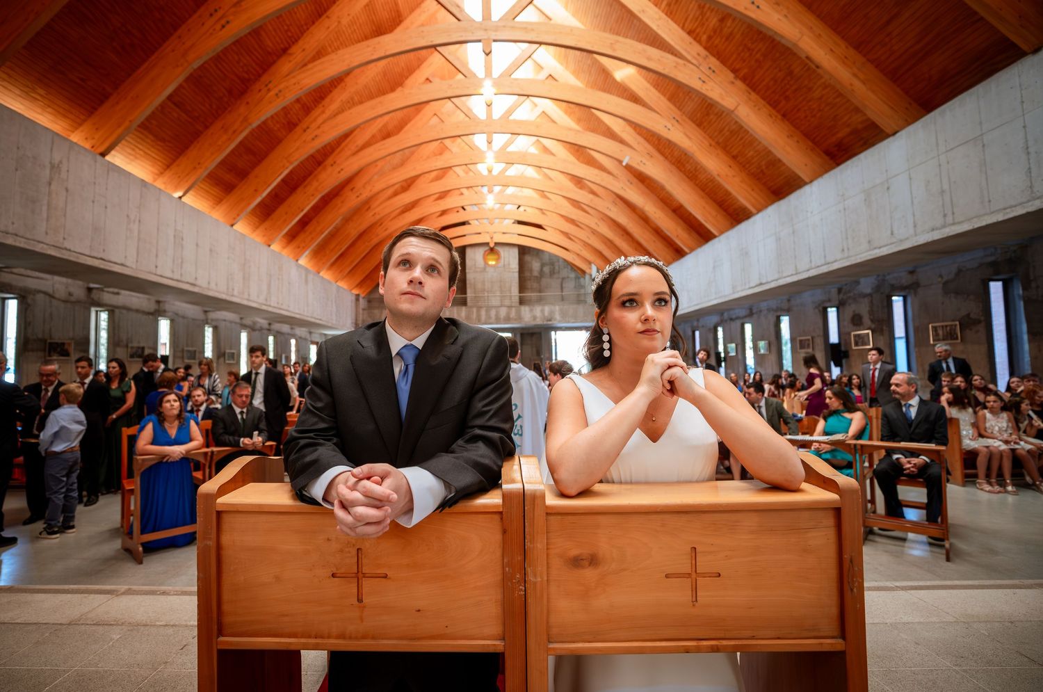 Bride and groom seated at wooden kneeler in a church with arched wooden ceiling and full pews.