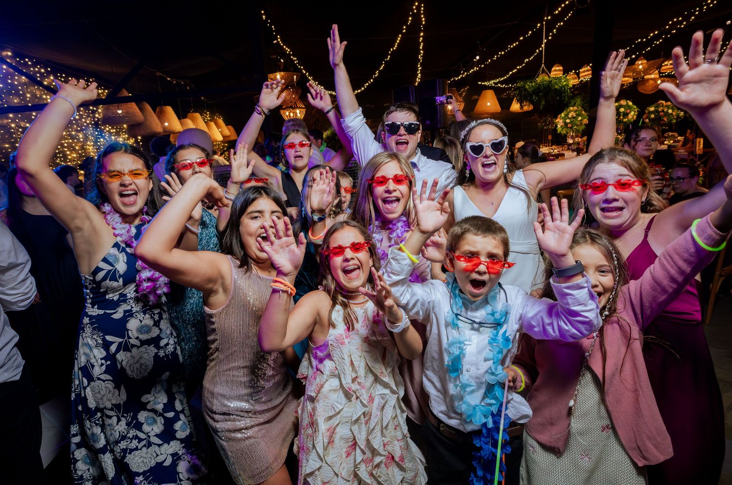 Bride and guests wearing red heart sunglasses celebrate with hands raised at wedding party.