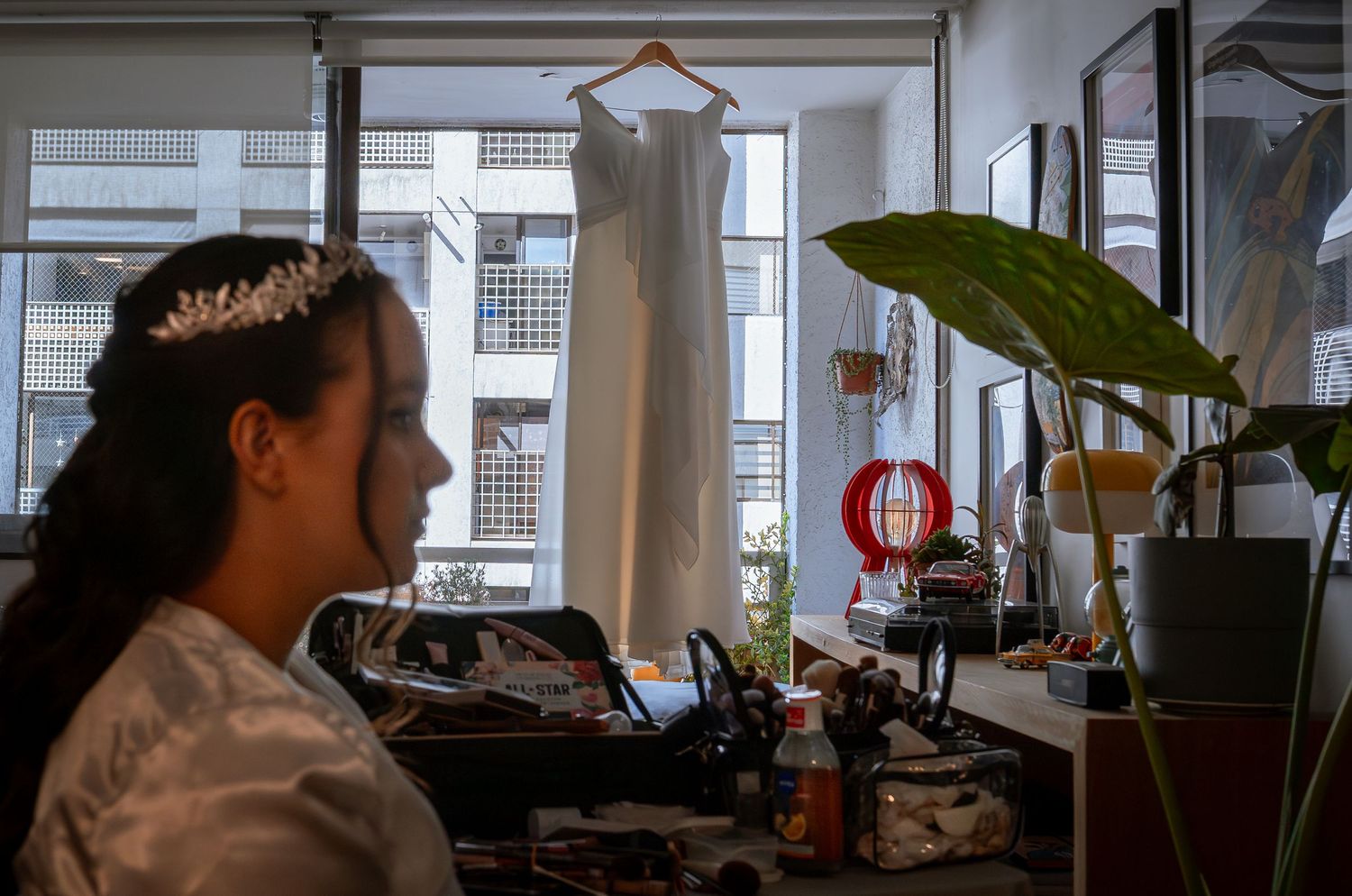 Bride with tiara sits at vanity with wedding dress hanging in background.