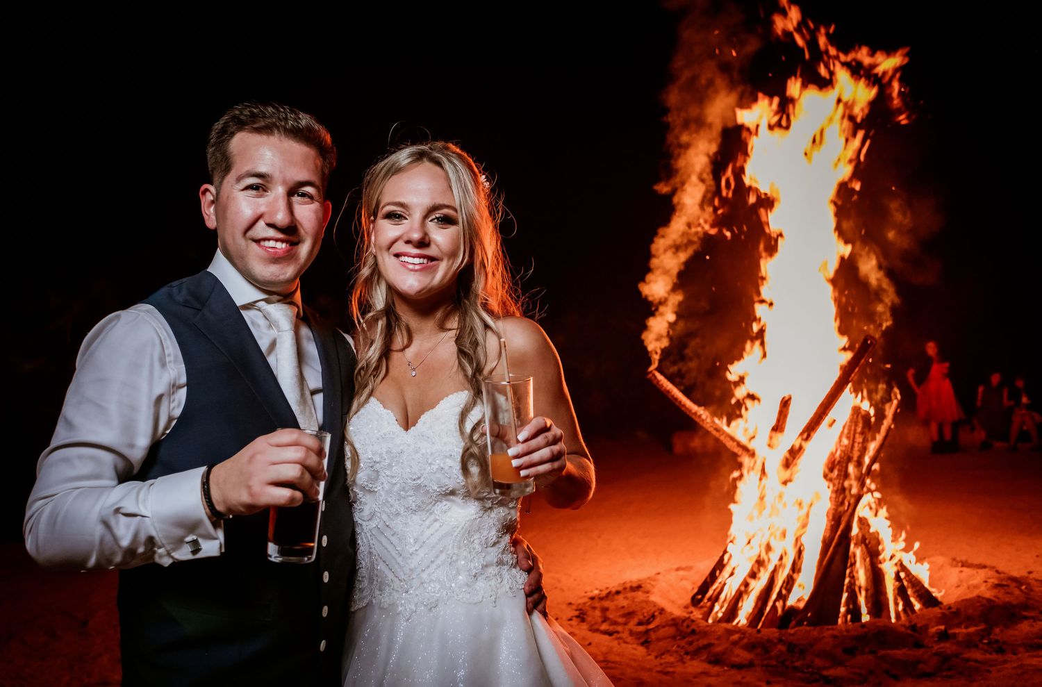 Bride and groom smiling together in front of a large bonfire at their outdoor wedding reception at night.