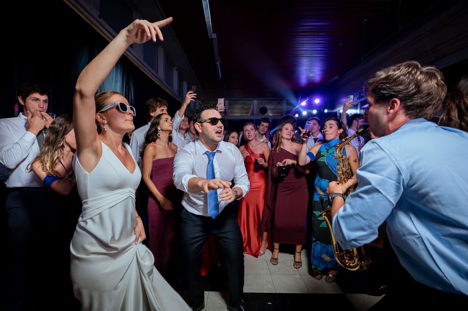 Bride and groom dancing energetically with saxophonist performing at wedding reception party.