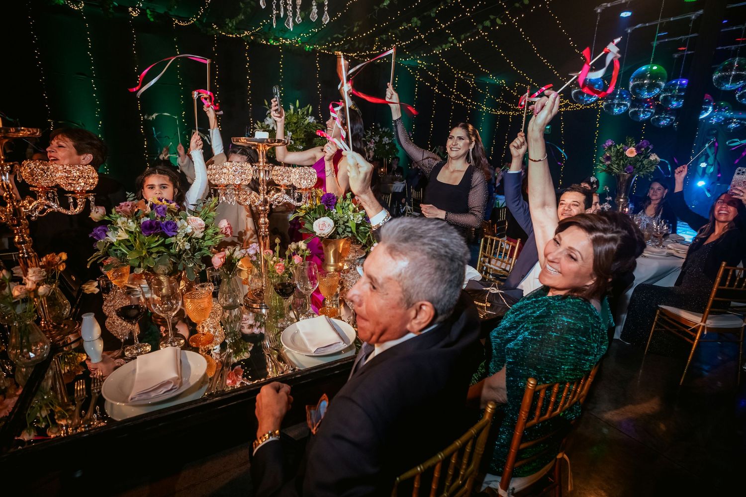 Guests celebrate at an elegant wedding reception with colorful streamers, lush floral centerpieces, and festive lighting.