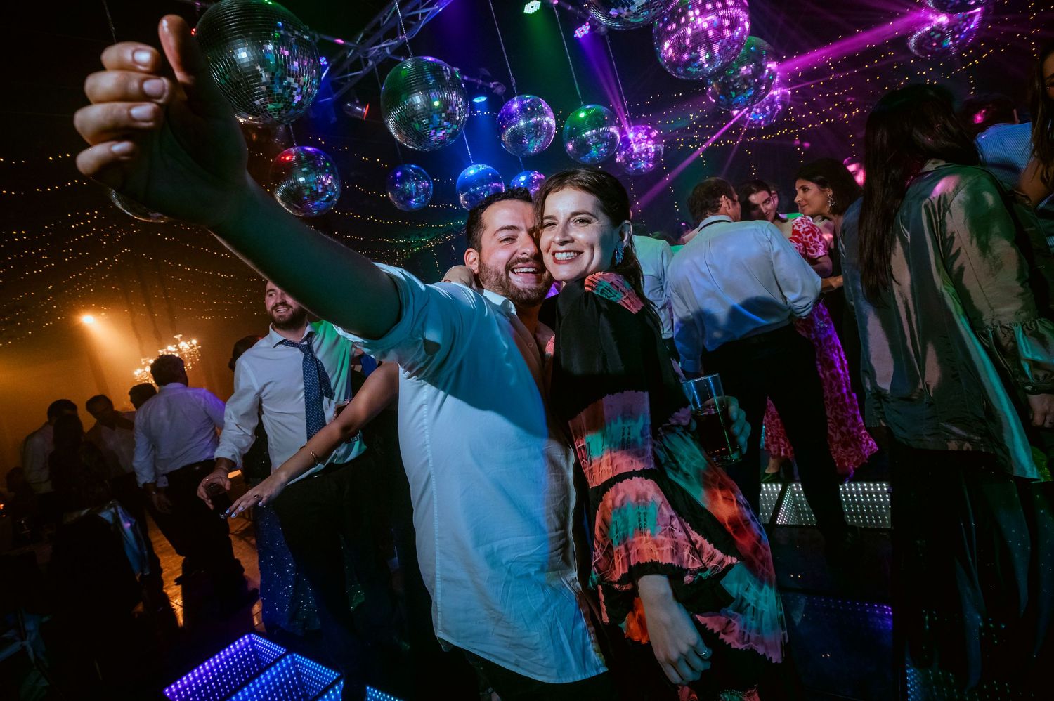 Two people taking a selfie at a vibrant nightclub party with disco balls and colorful lights in the background.