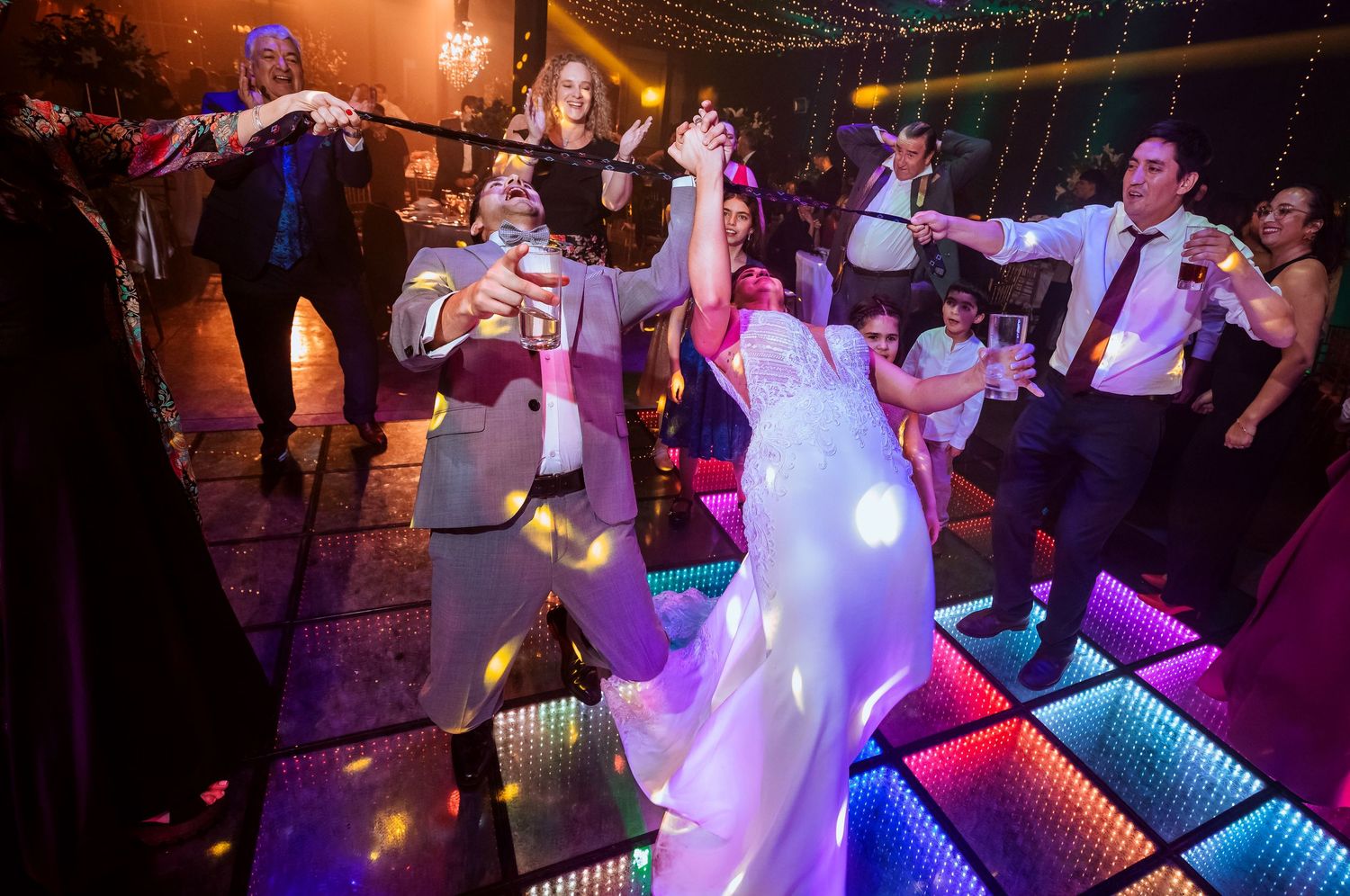 Bride and groom dancing joyfully on colorful LED dance floor surrounded by wedding guests at elegant reception.